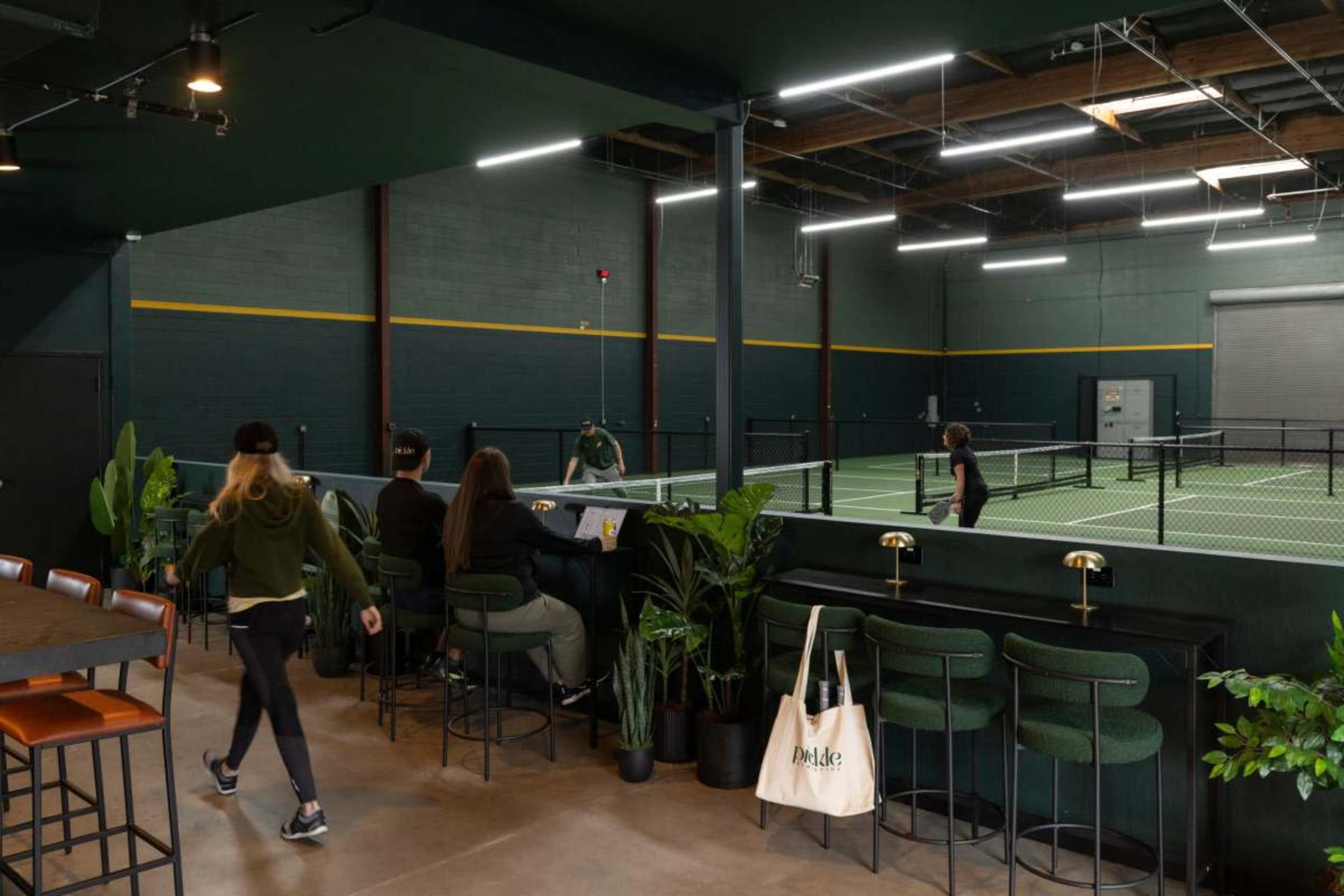 The image shows a modern indoor tennis facility with greenery, seating at a bar, and players on the courts in the background.