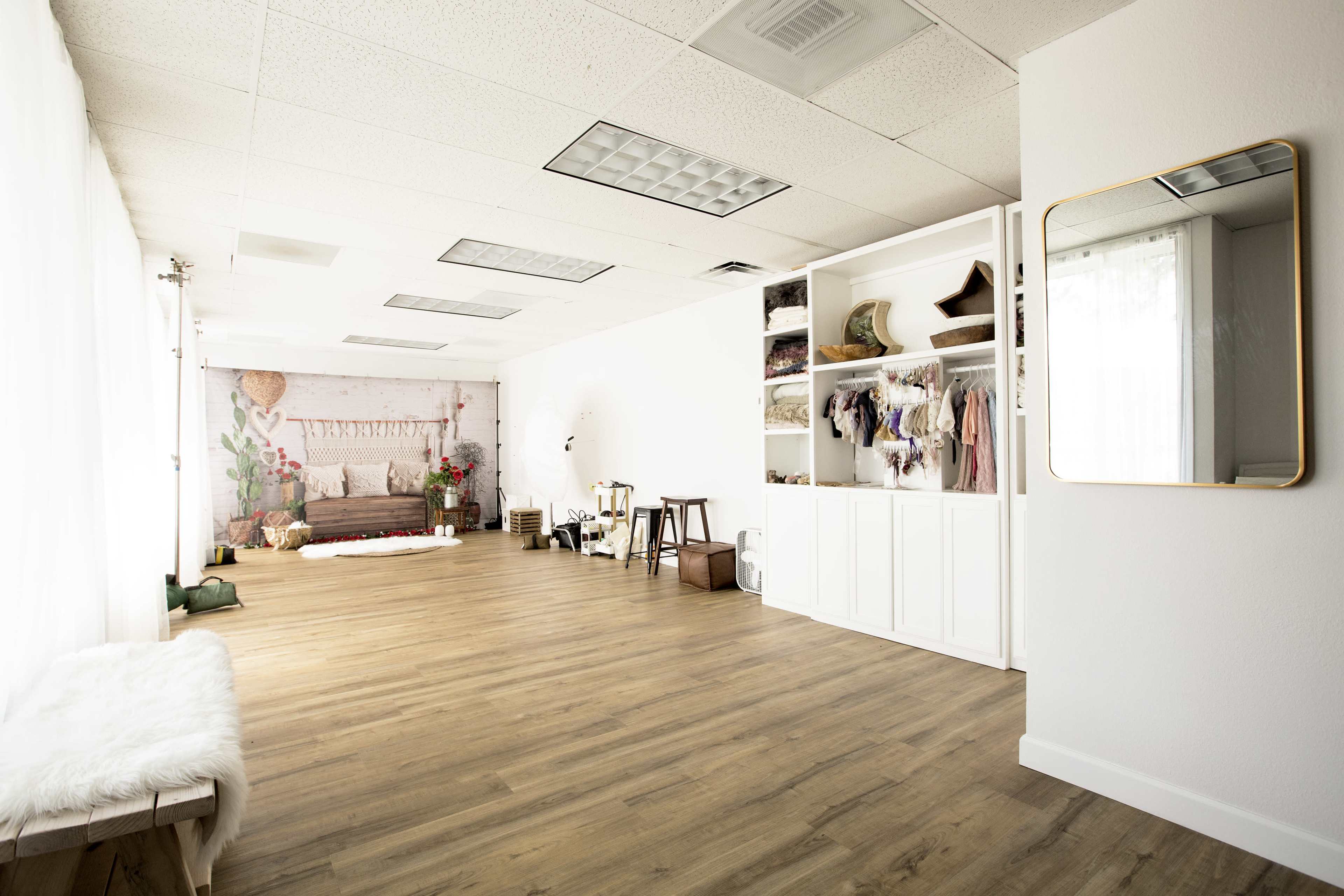The image depicts a spacious, well-lit studio with a wooden floor, showcasing a seating area, storage shelves, and a large mirror along the wall.
