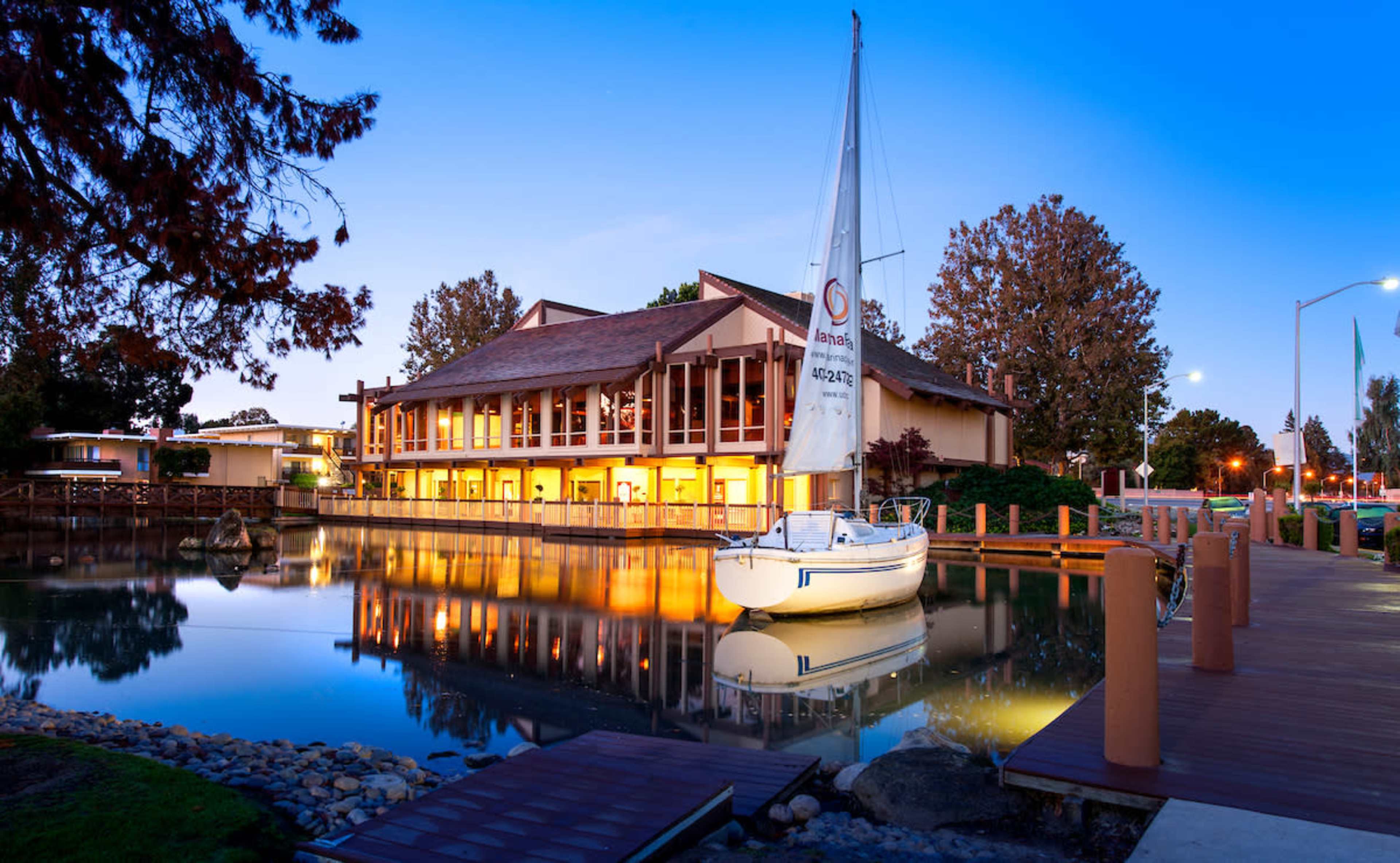 A white sailboat is moored beside a wooden dock in front of a multi-story building reflecting on the calm water at dusk.