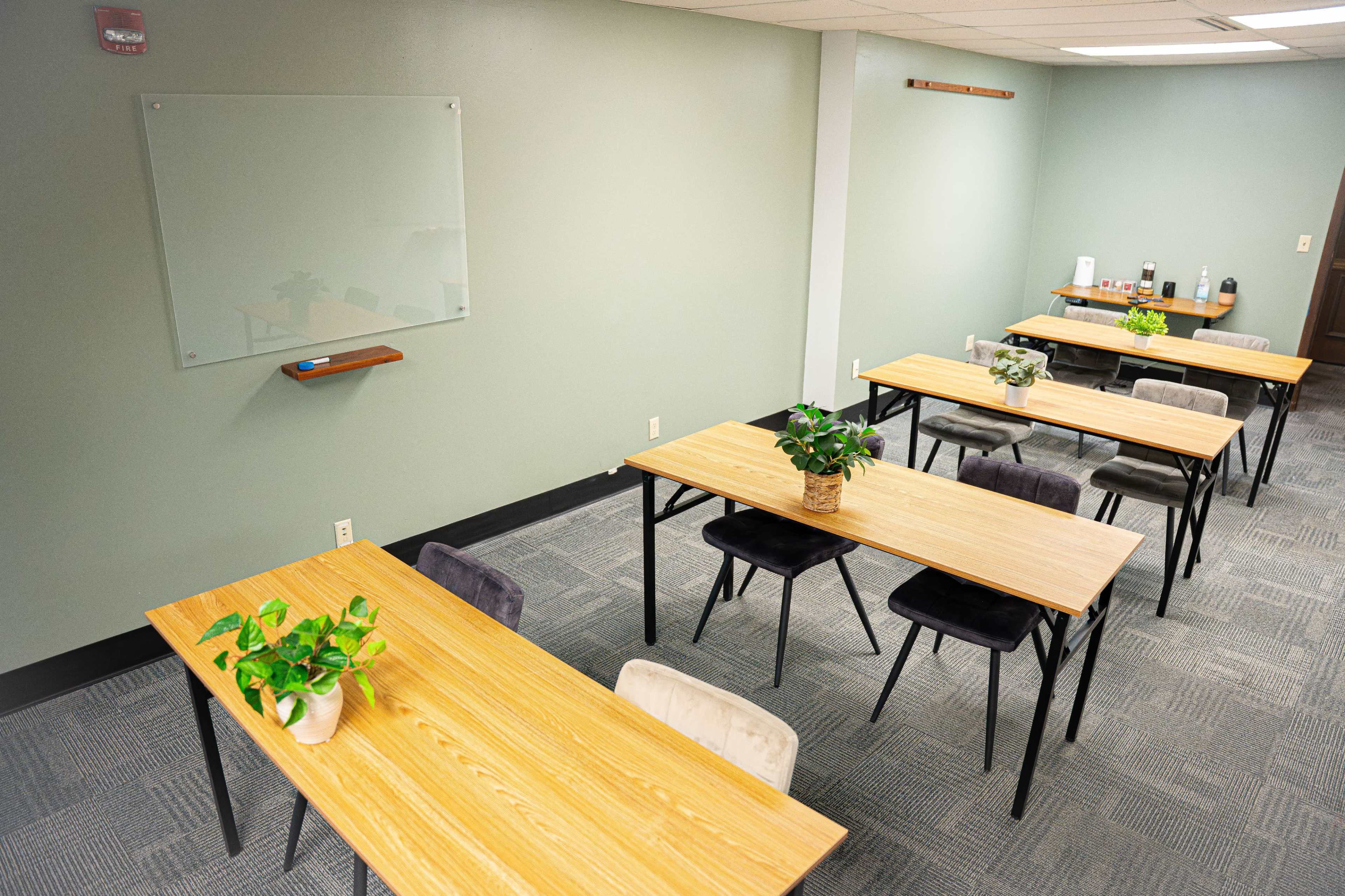 The image shows a classroom setting with several wooden tables arranged neatly, chairs beside them, and green plants on the tables, with a whiteboard mounted on the wall.