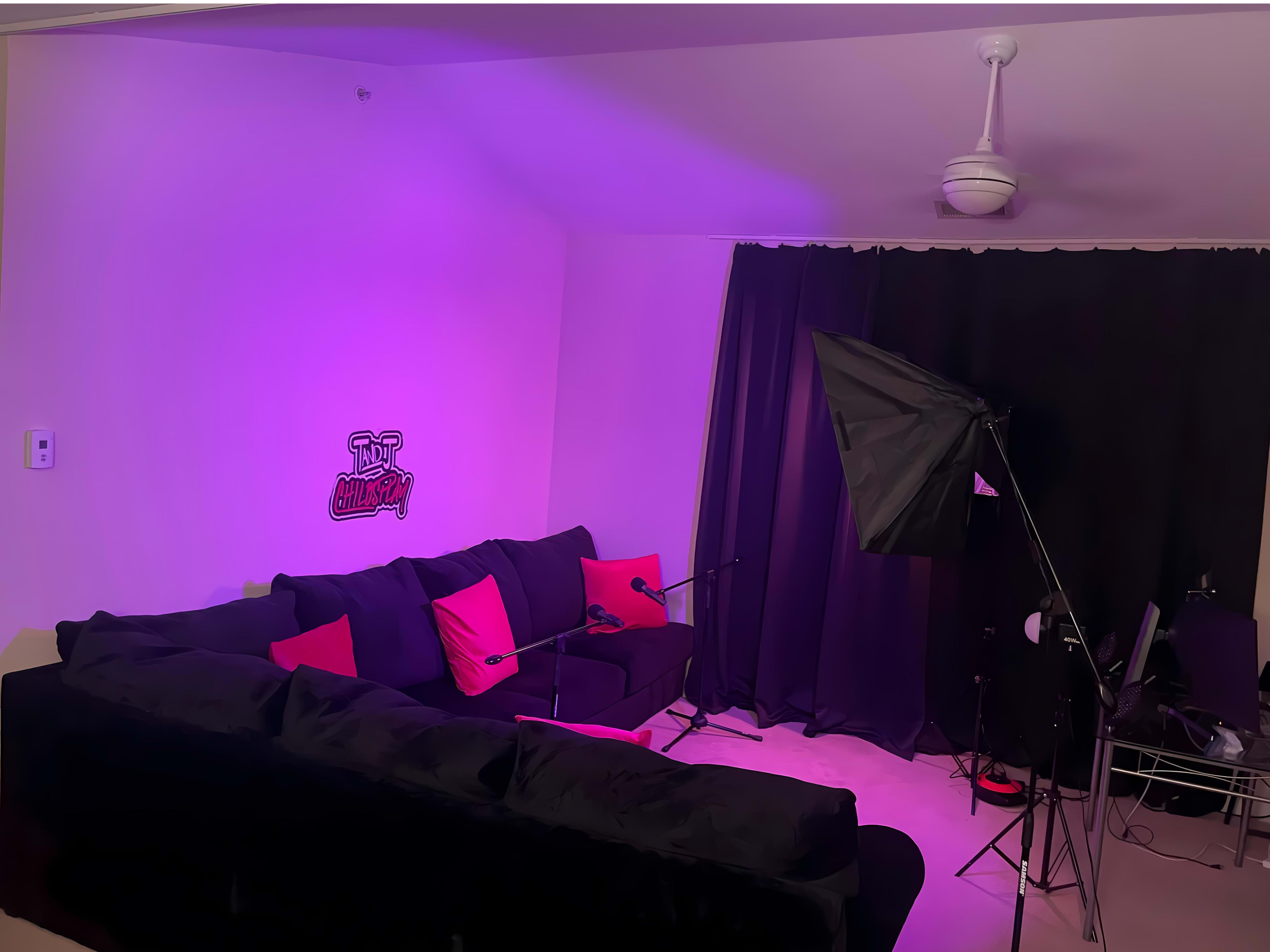A living room features a large black couch adorned with red cushions, against a backdrop of purple lighting and black curtains, with a microphone setup and a camera on a stand.