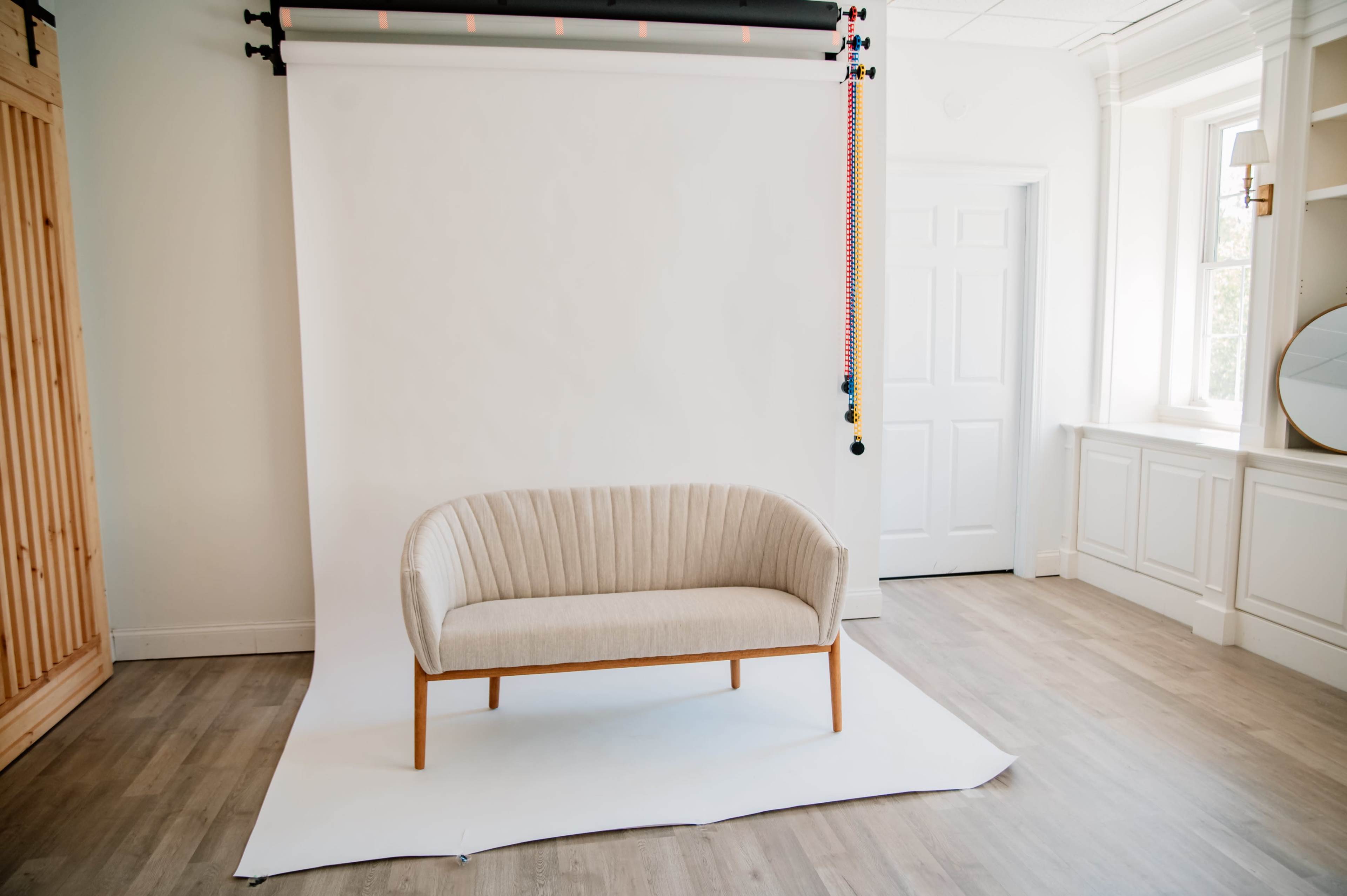 A beige upholstered couch with wood legs is centered in a bright, minimalistic room with white walls and hardwood flooring.