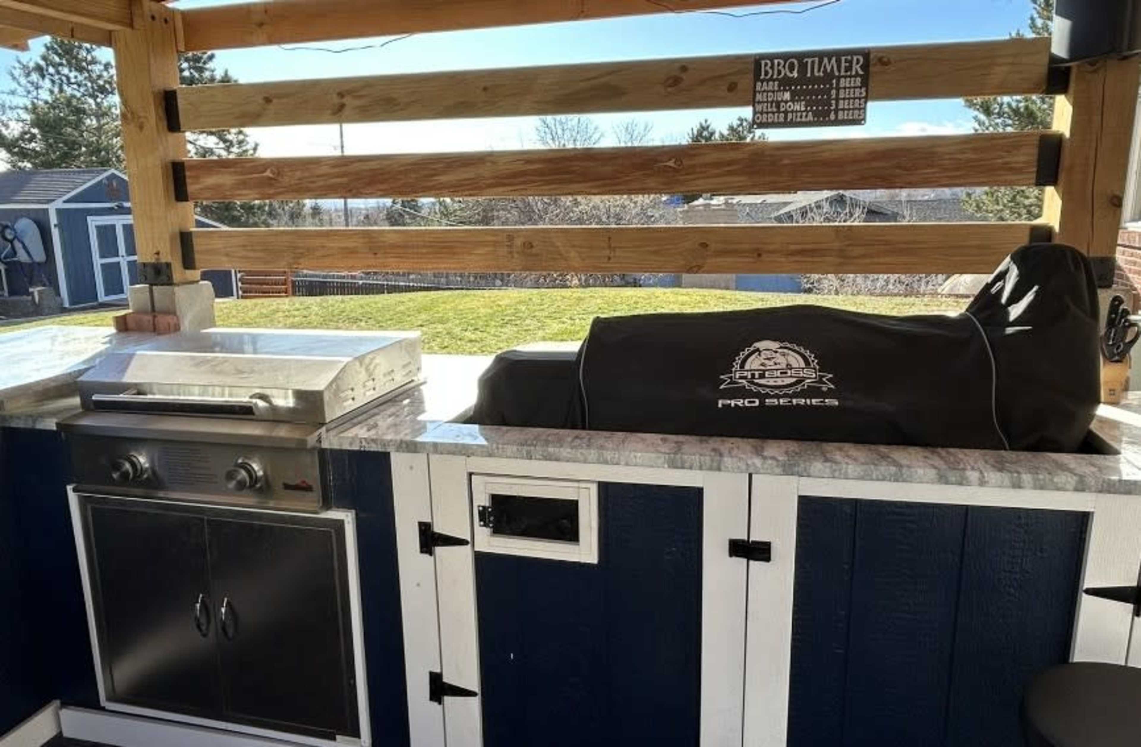 The image shows an outdoor cooking area featuring a stainless steel grill and a covered storage space, with a backdrop of a grassy yard and distant trees.