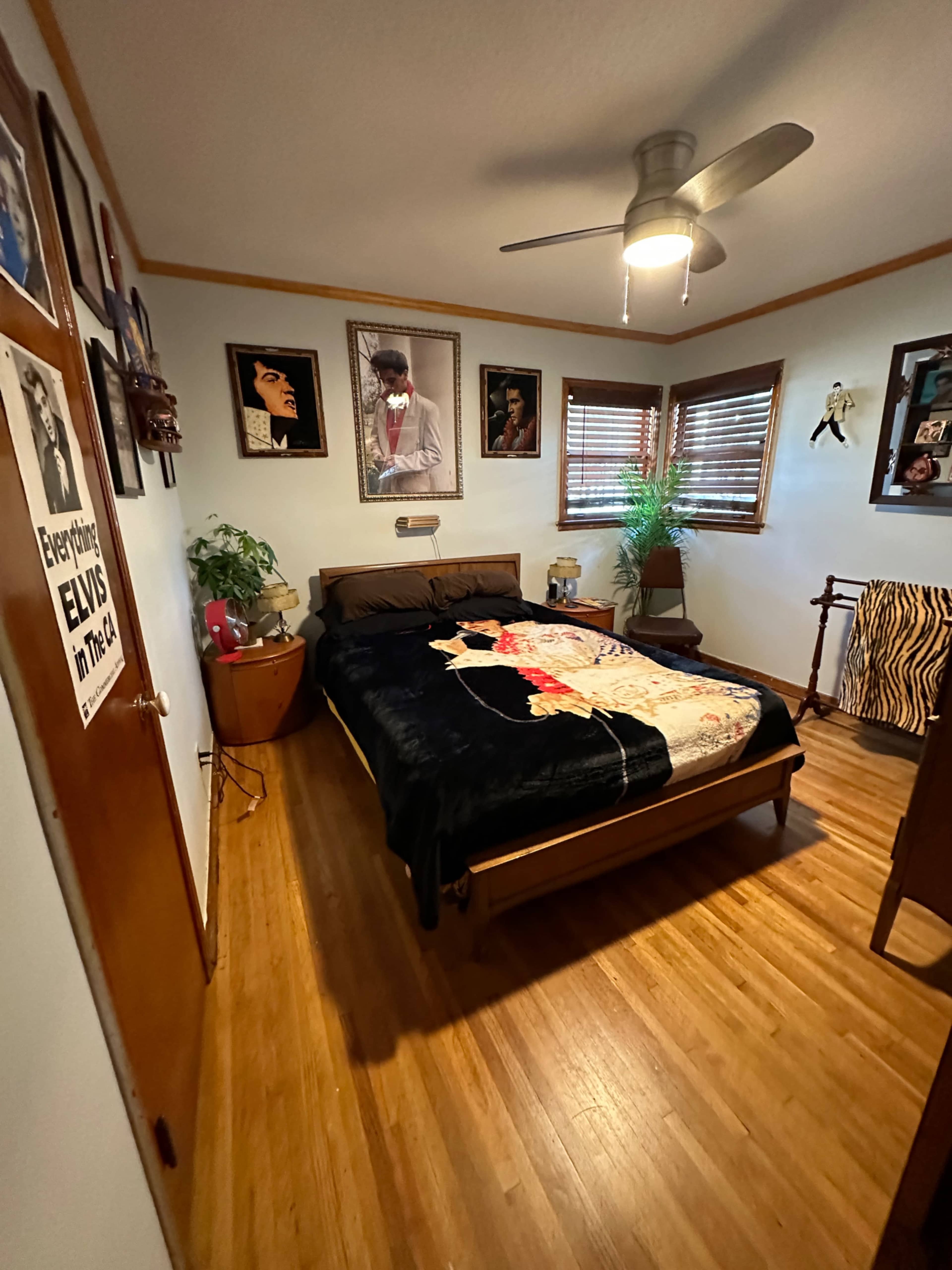 A bedroom with a bed covered in a black blanket, wooden flooring, and framed artworks on the walls, including a prominent poster of Elvis Presley.
