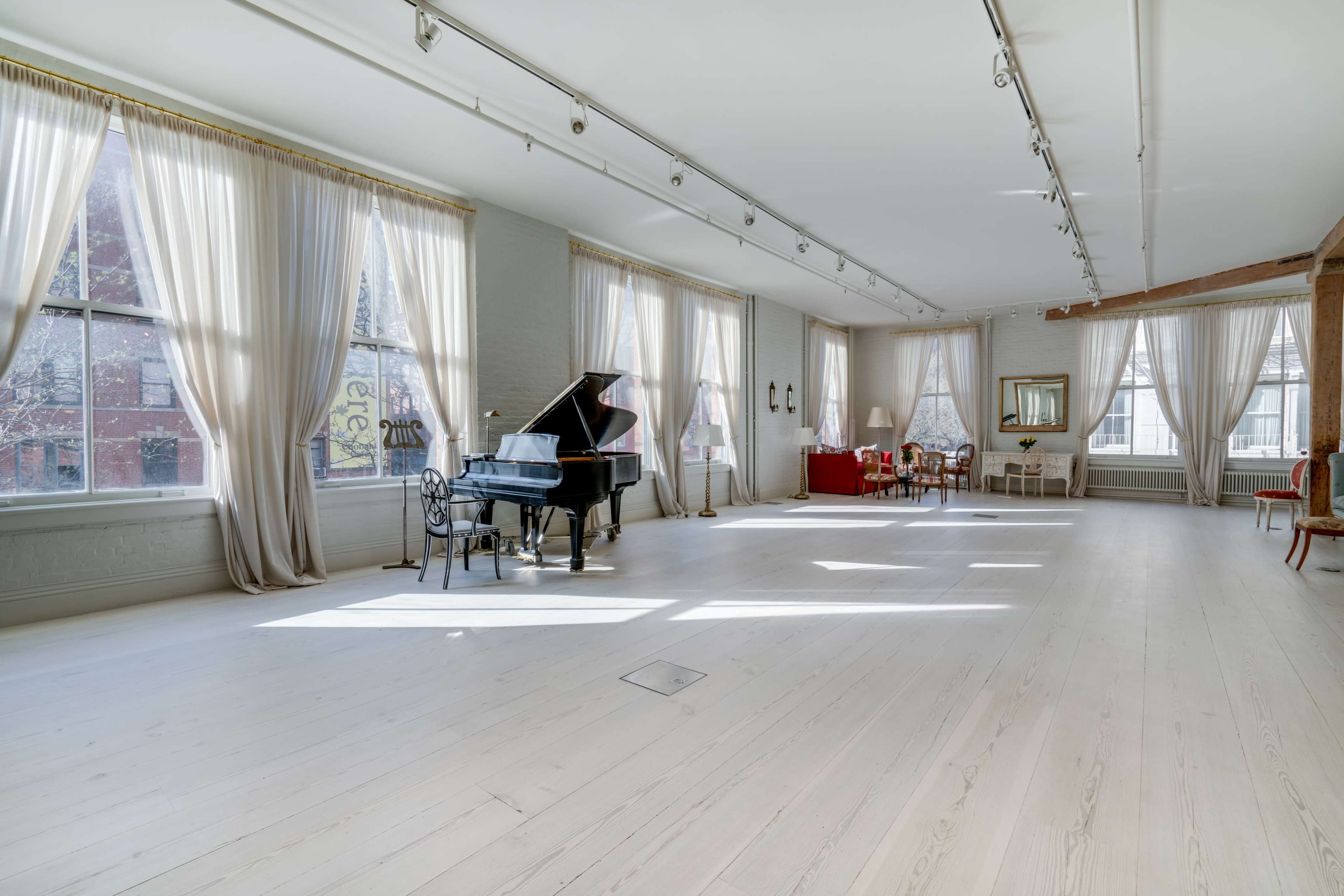 A spacious room features a grand piano, multiple large windows with drapes, and a seating area with elegant furniture.