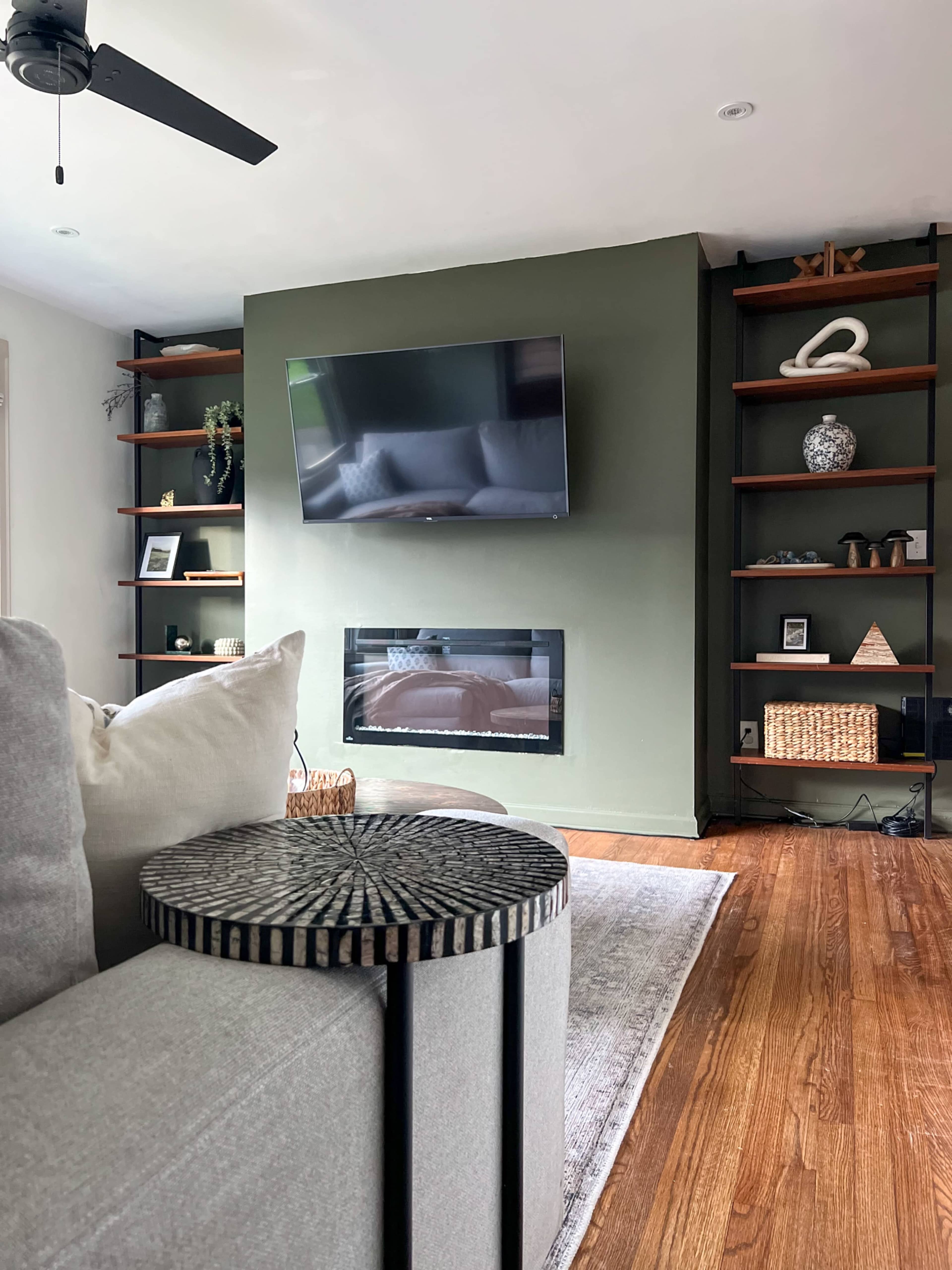 The image shows a modern living room featuring a green accent wall with a mounted television above a wall-mounted fireplace, flanked by wooden shelves holding decorative items.