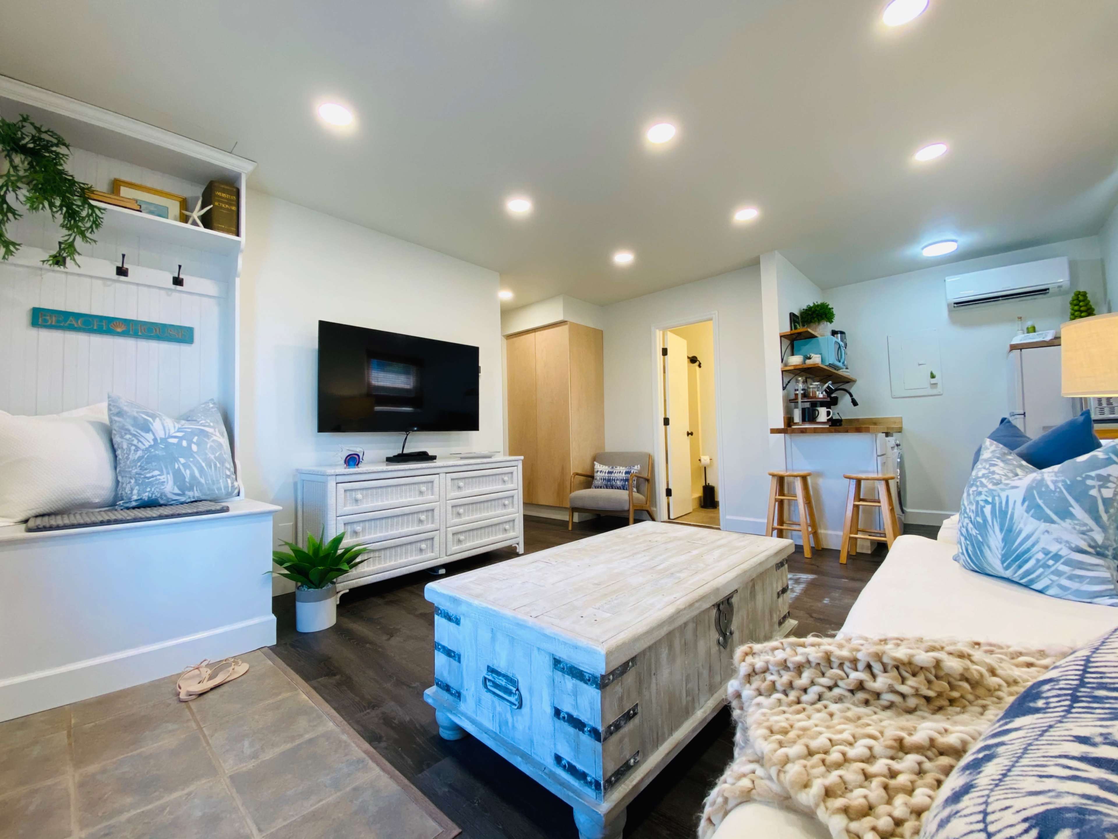 A modern living room features a white sofa, a wooden coffee table, a flat-screen TV on a console, and a cozy reading nook with decorative pillows.