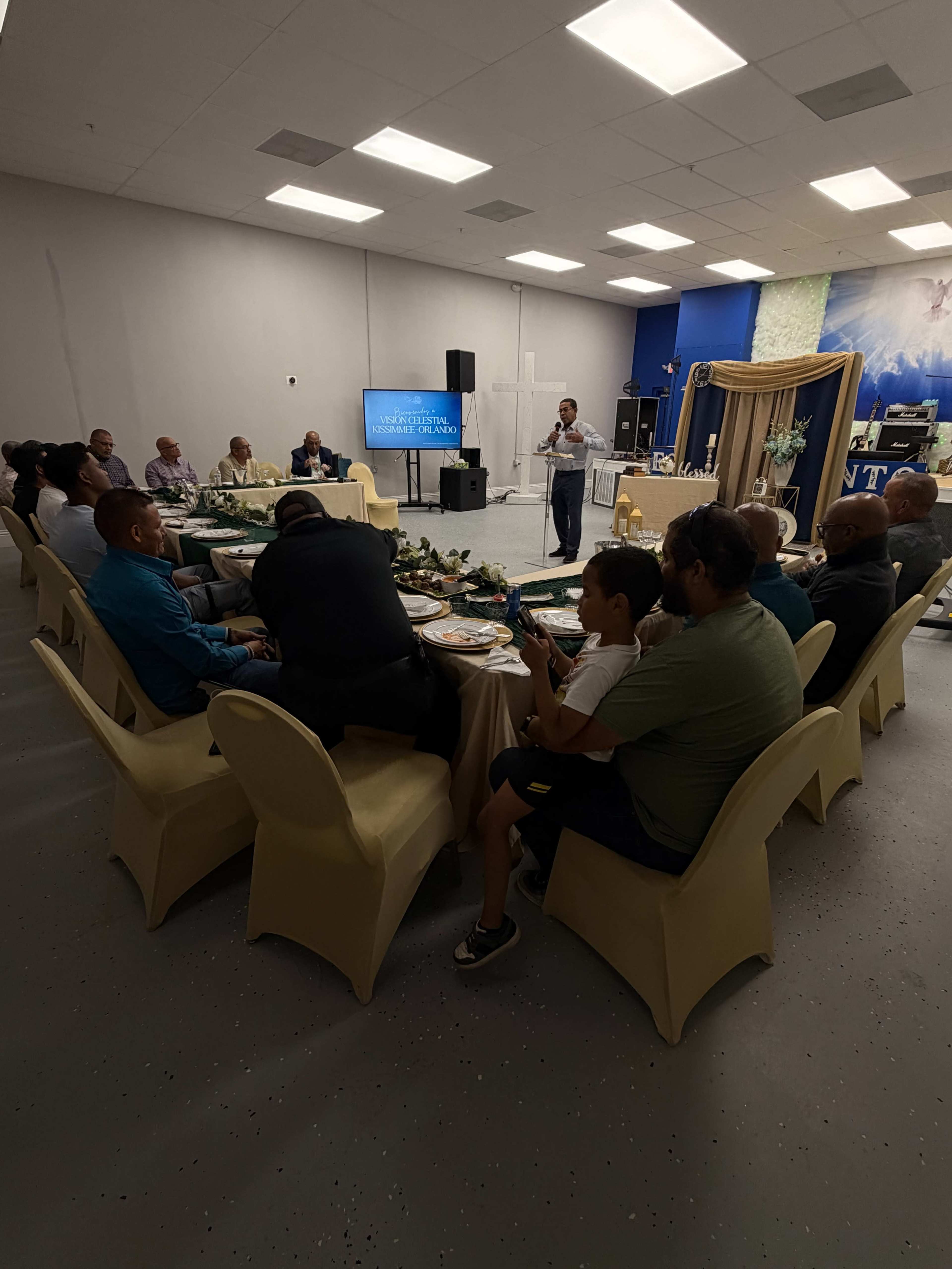 A group of people is seated around a circular table in a banquet hall, with someone speaking in front of a presentation screen.