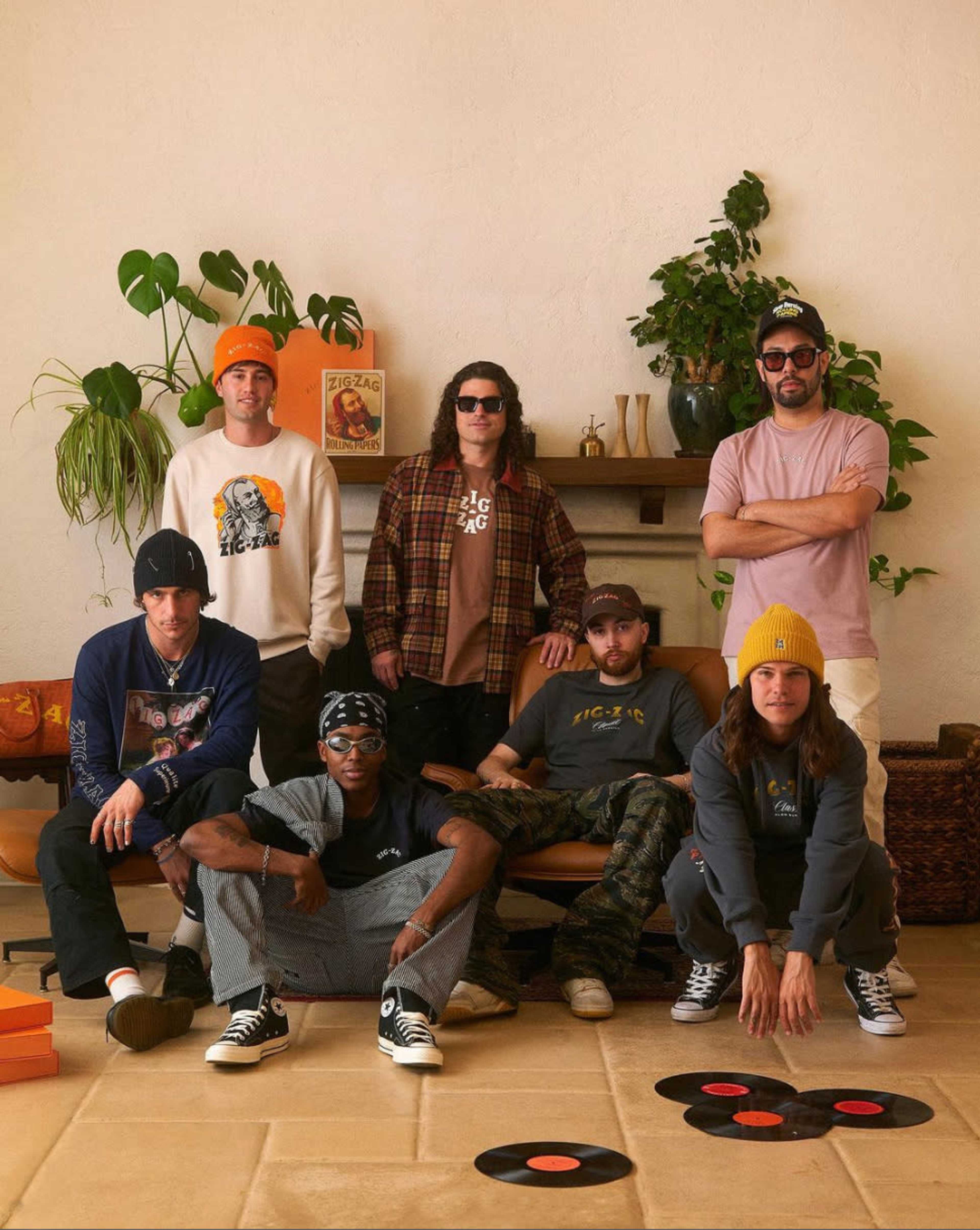A group of seven people poses in a room with plants and a retro record player, wearing casual streetwear and accessories.