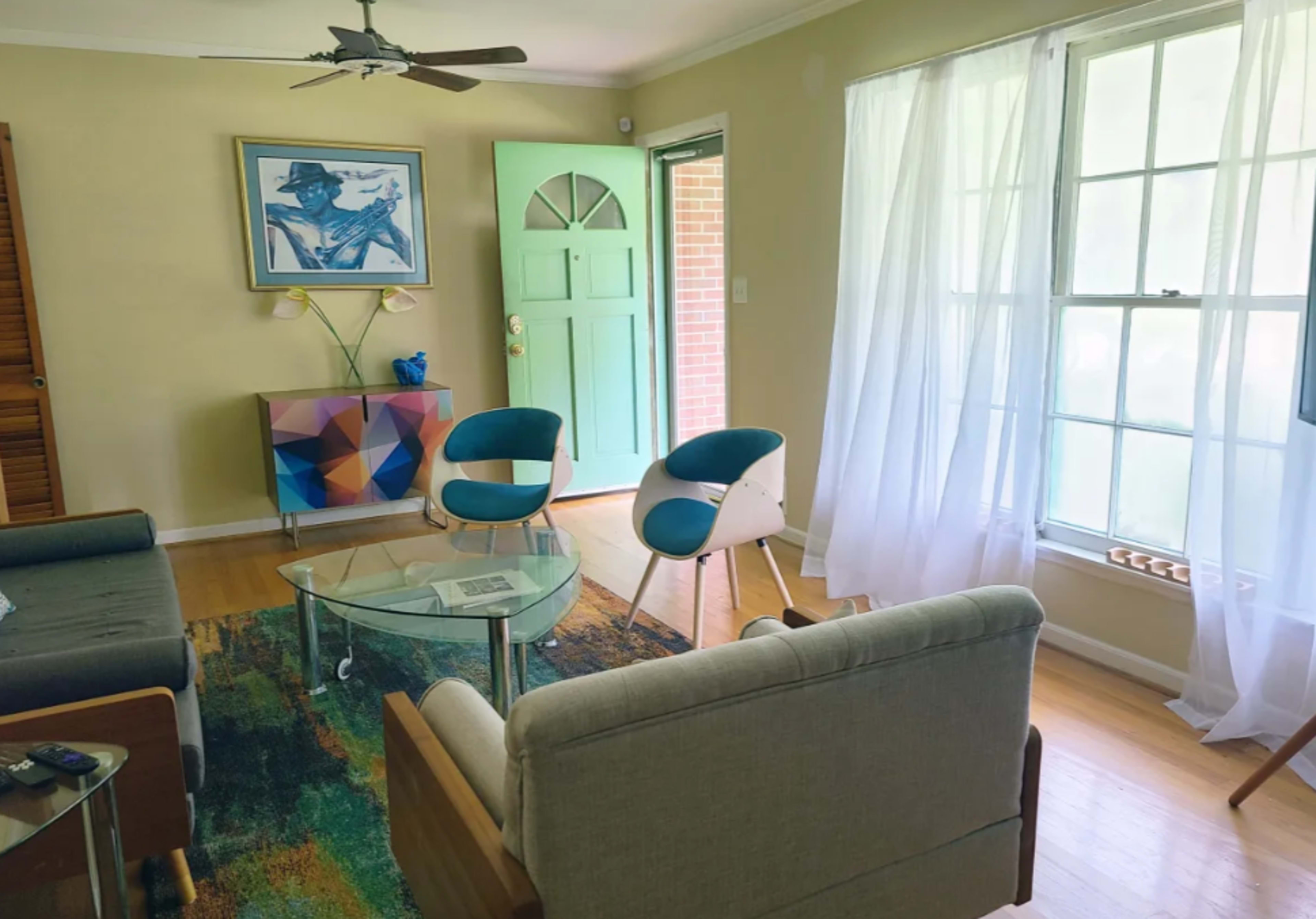 A living room features a glass coffee table, two blue chairs, and a colorful geometric side table near a large window with sheer curtains.