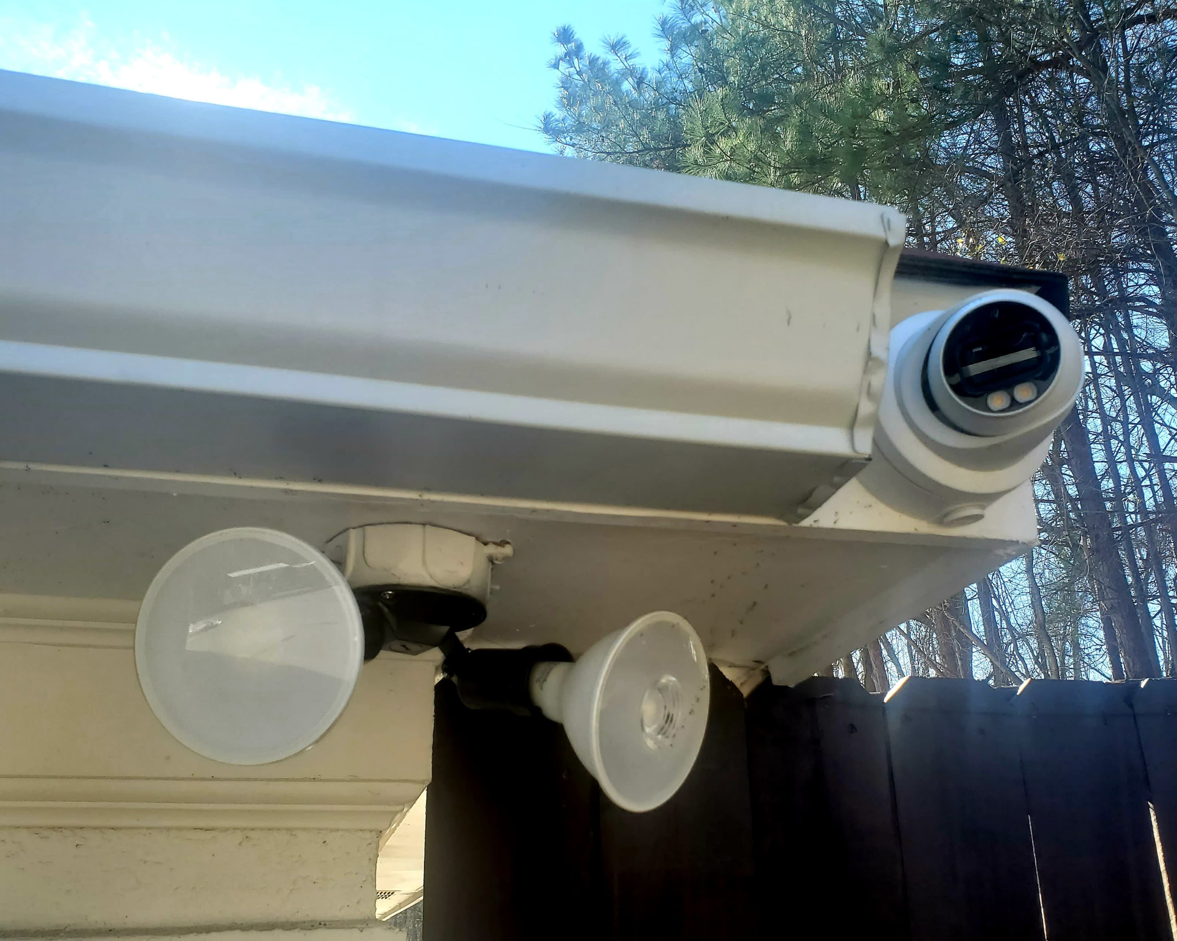 Two outdoor lights mounted on a house's eaves, with one light featuring a circular socket and the other a clear plastic covering.