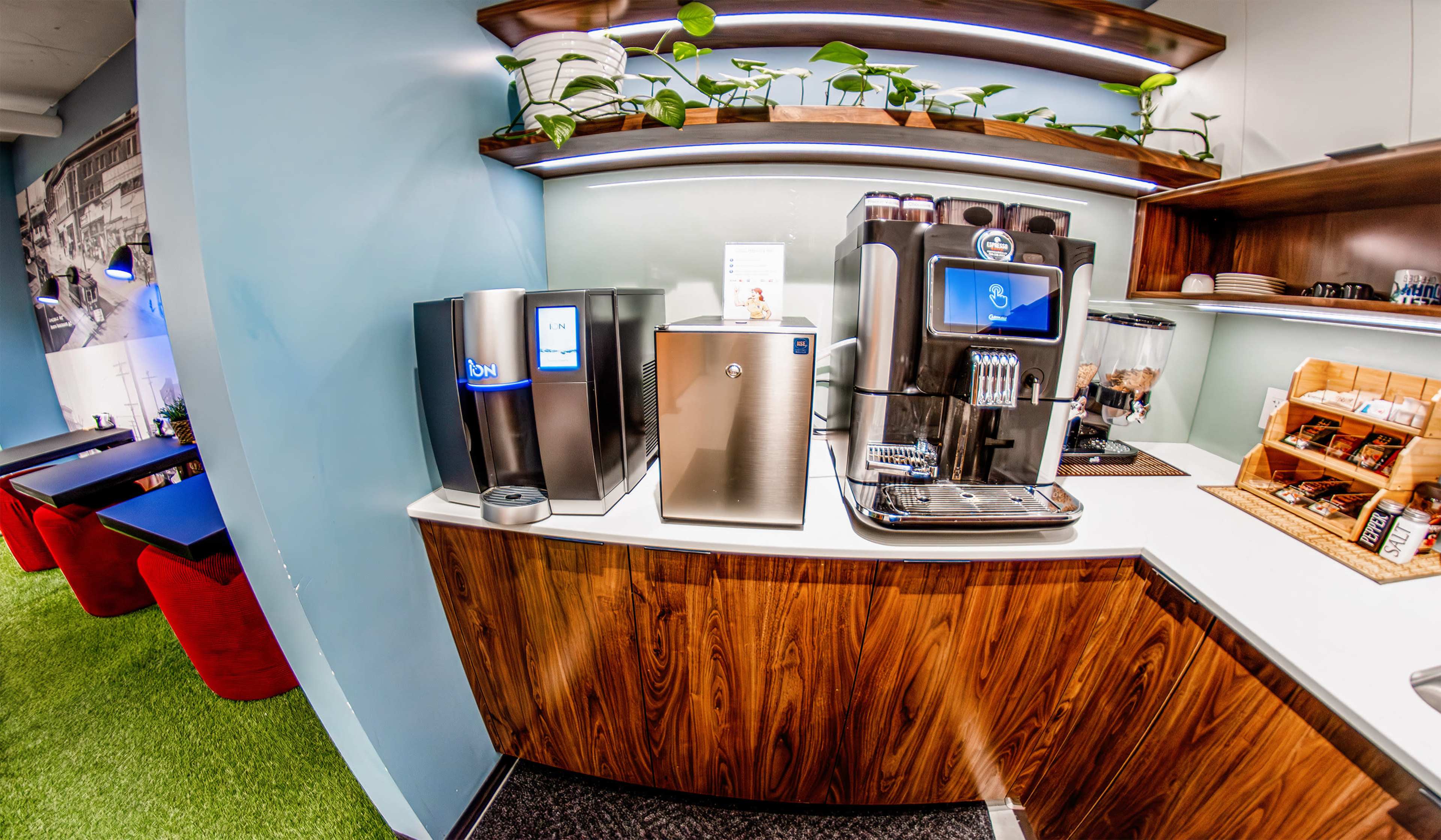 The image shows a modern kitchenette with a coffee machine, water cooler, and shelves adorned with plants, all positioned against a stylish wall.