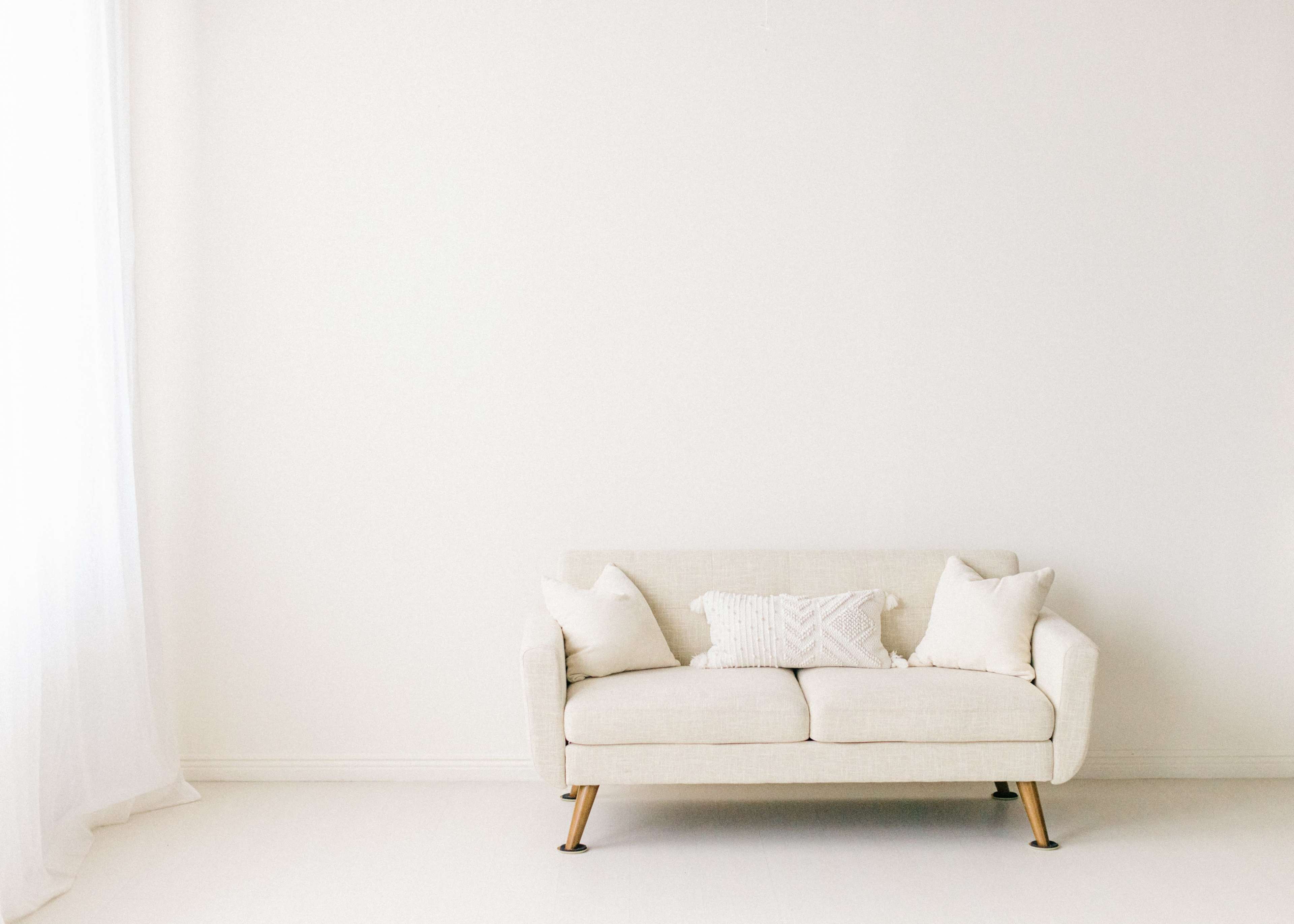 A light-colored sofa with two cushions sits against a plain white wall in a bright, minimalistic room.