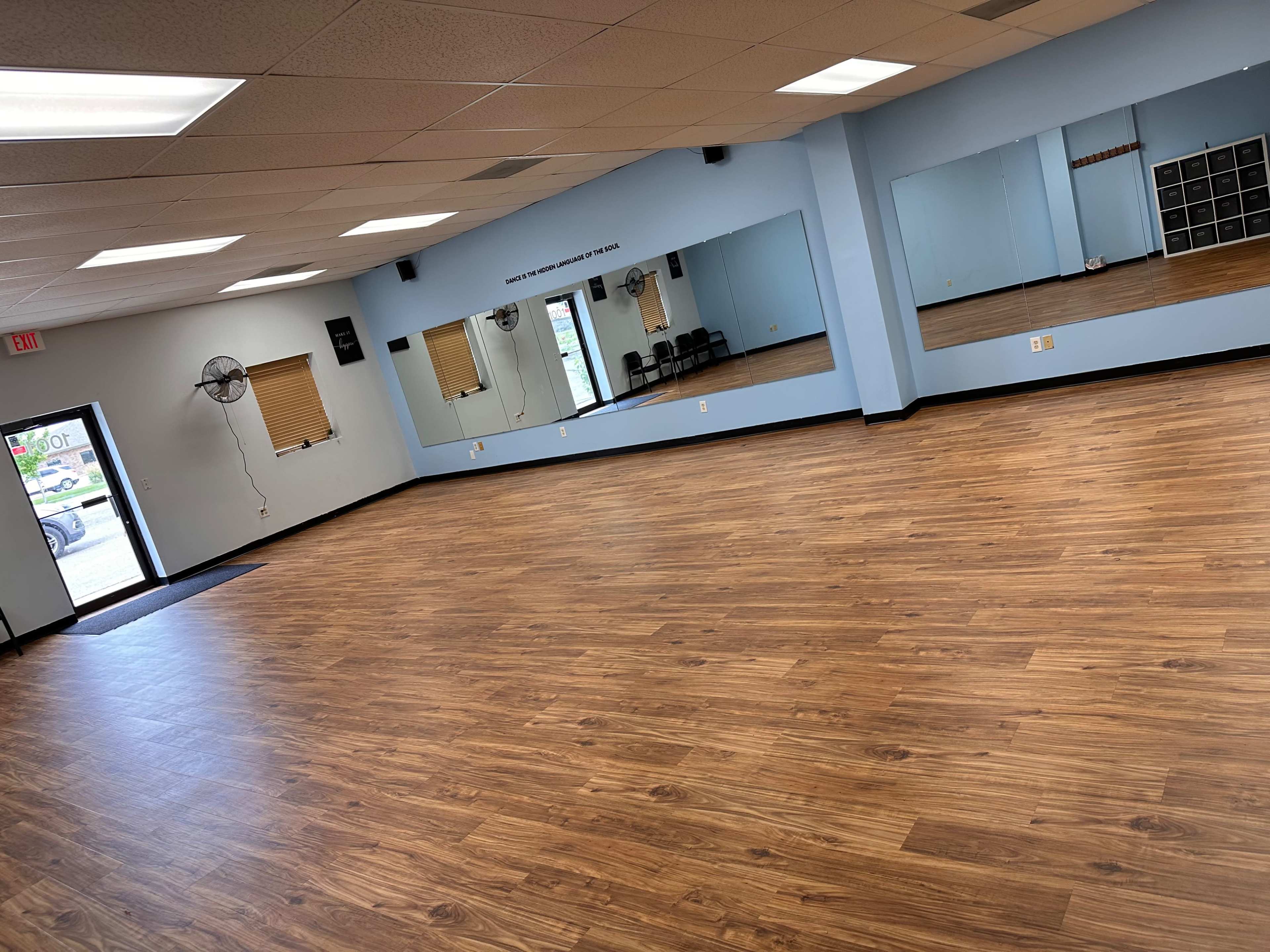The image shows an empty dance studio with wooden flooring, blue walls, mirrors on one side, and a door leading outside.