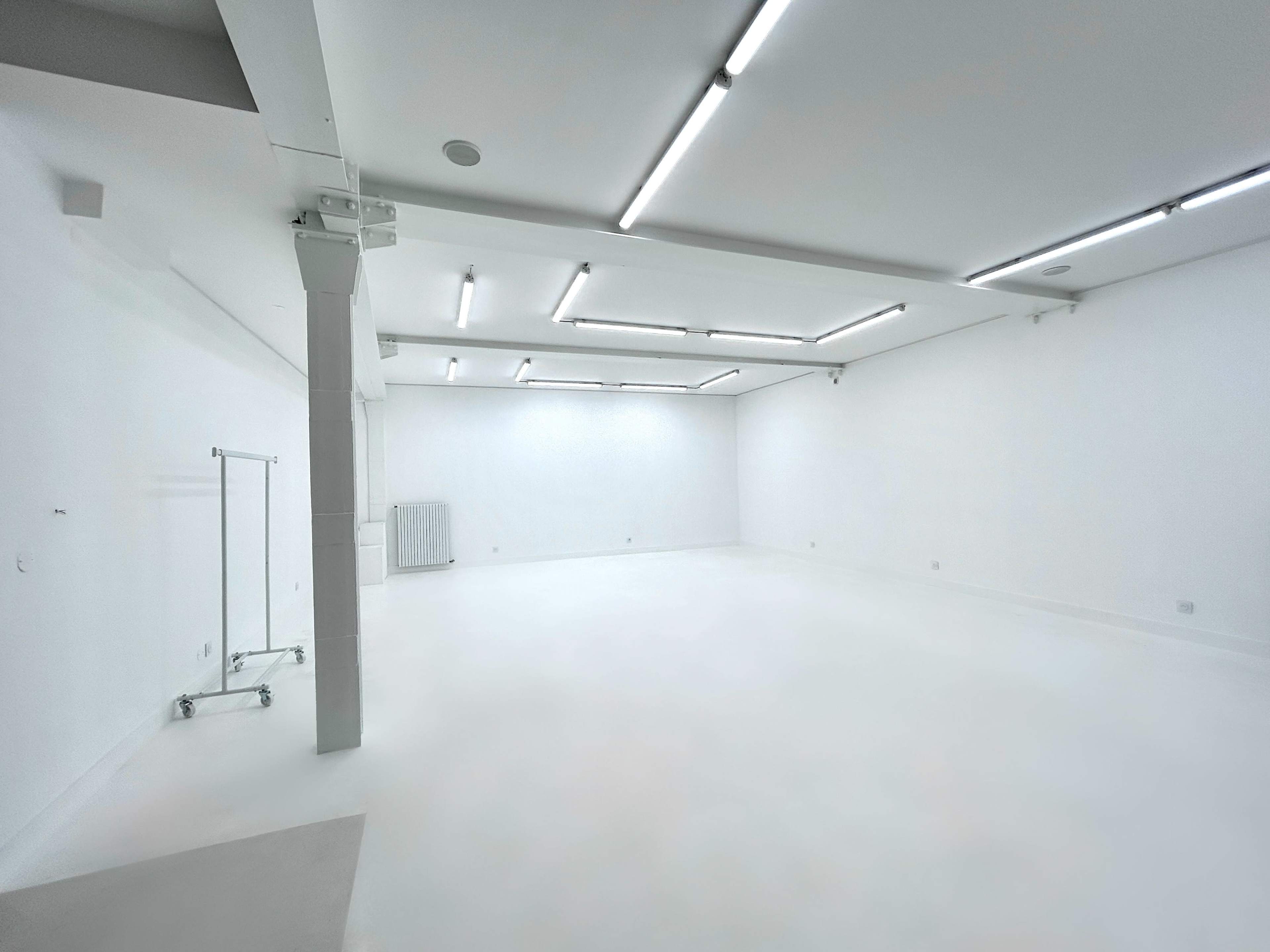 A spacious, minimalist room with white walls and a polished floor, featuring fluorescent ceiling lights and a clothing rack in one corner.