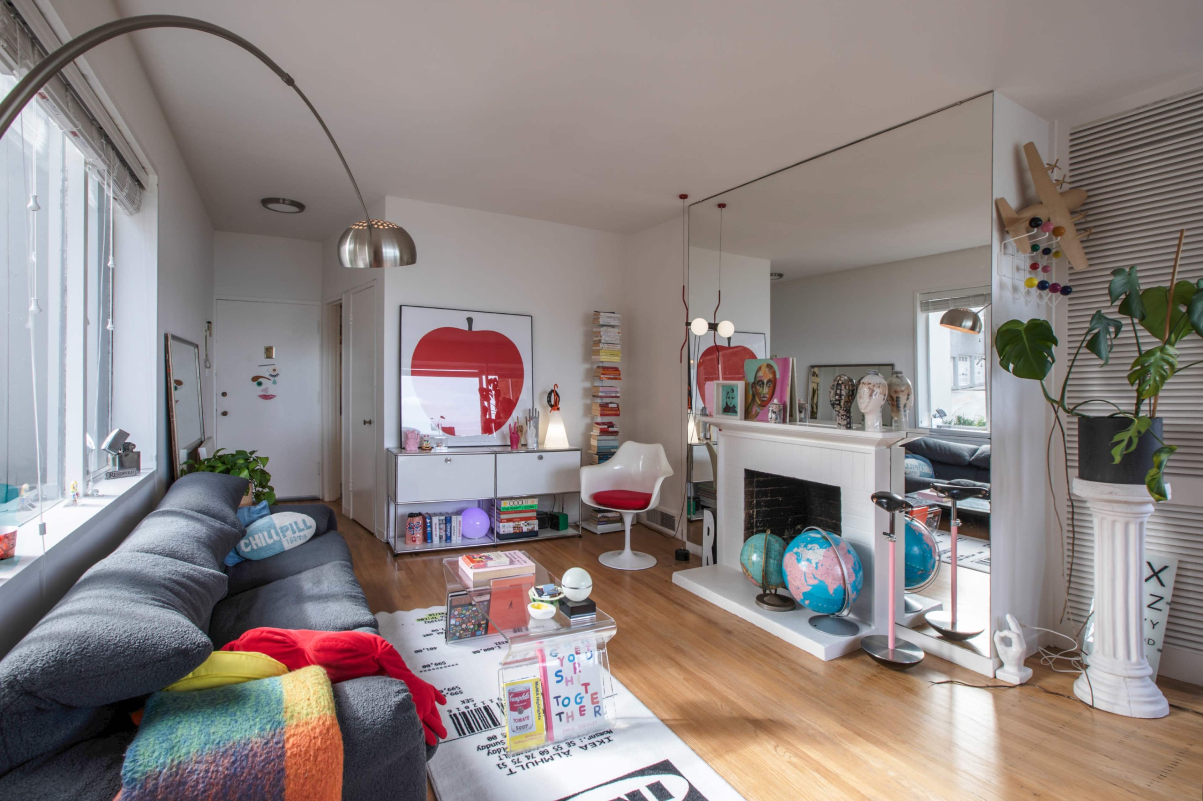 A bright living room features a gray sofa, a white shelving unit filled with books and decorative items, and a large mirror reflecting a cozy seating area with a globe and plants.