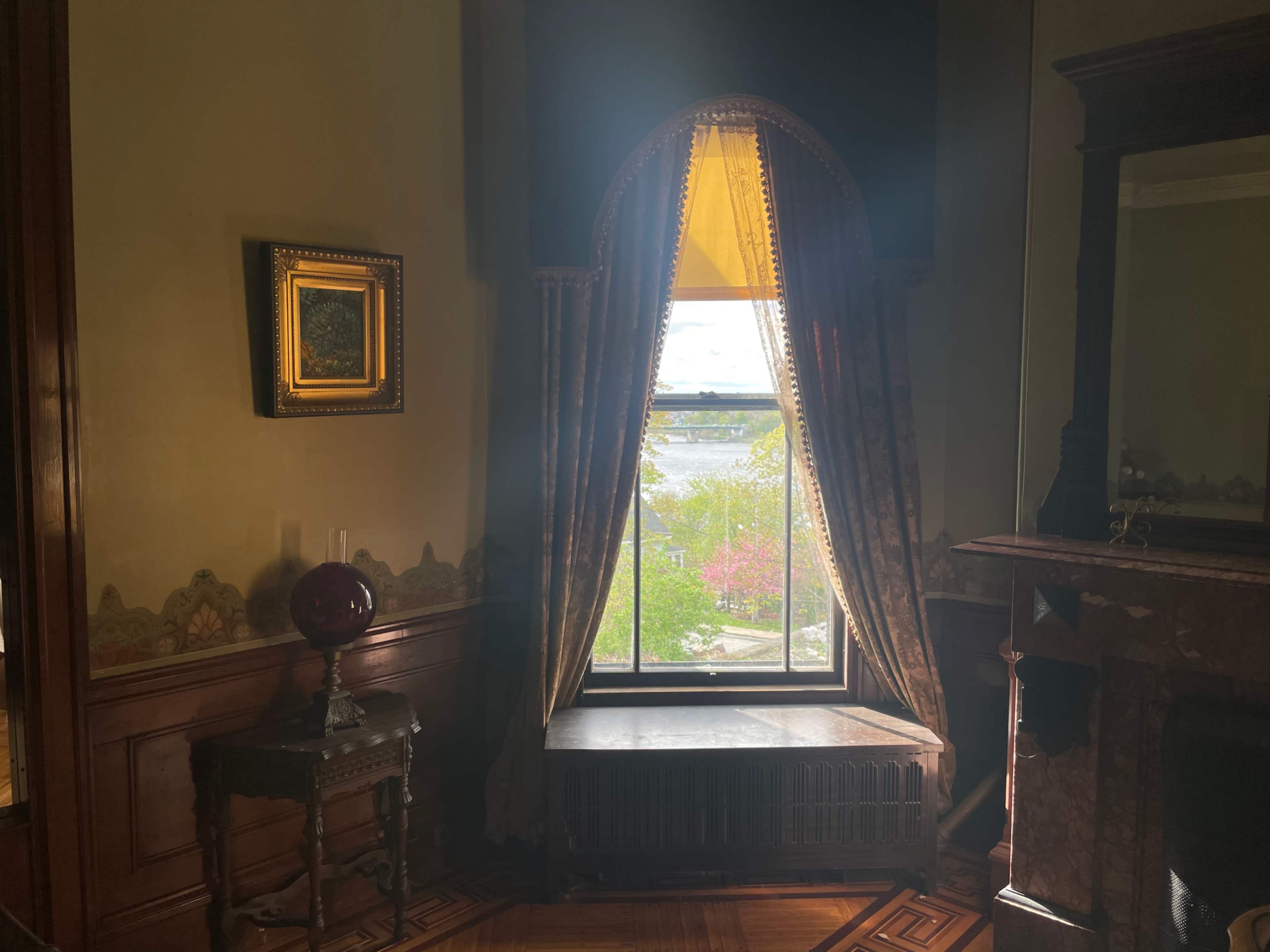 A window dressed with elegant curtains overlooks a garden with trees, framed by ornate wall moldings and a decorative fireplace in a room.