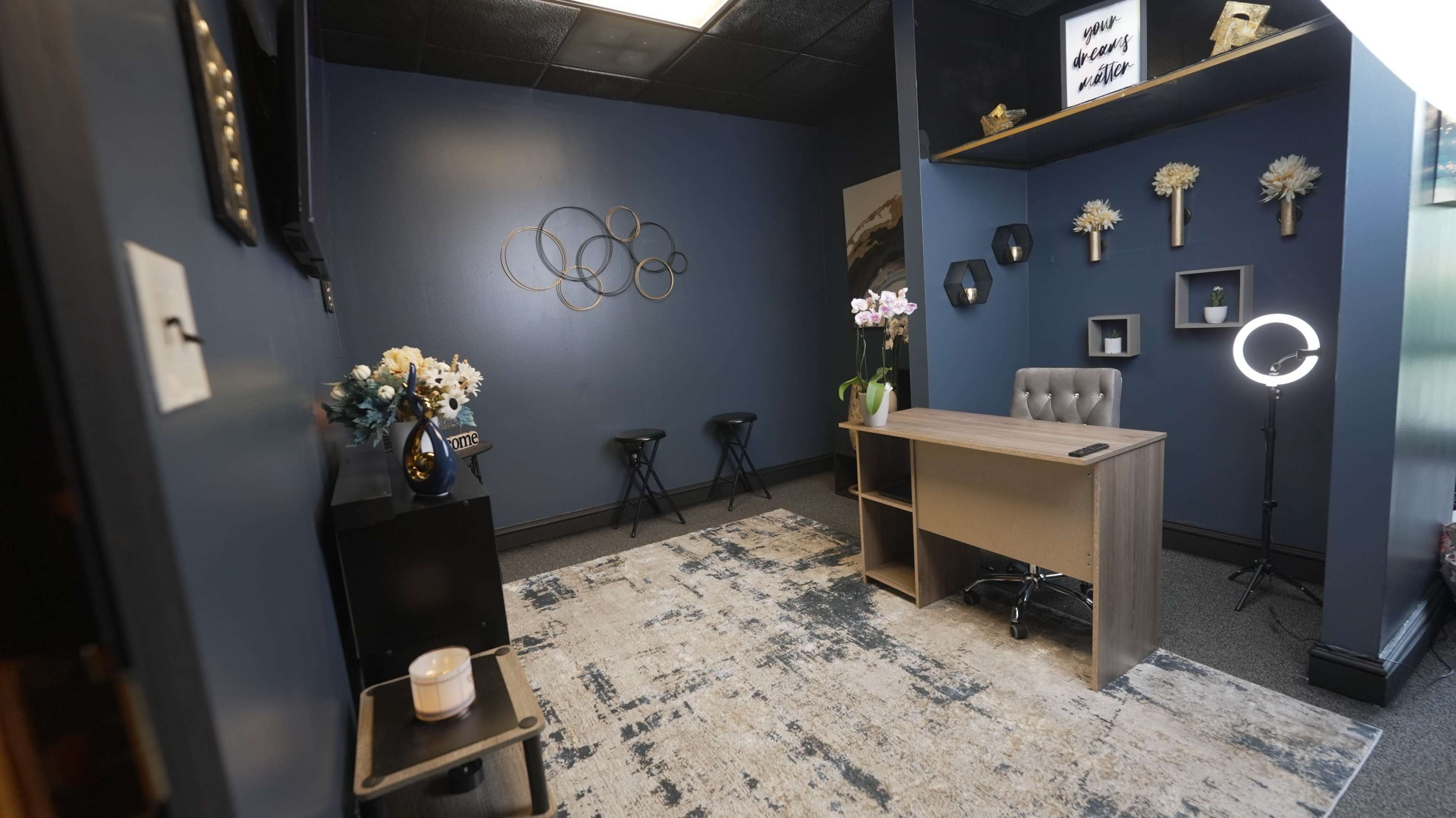 The image shows a small office space with dark blue walls, a wooden desk, a comfortable chair, decorative shelves, and a rug on the floor.
