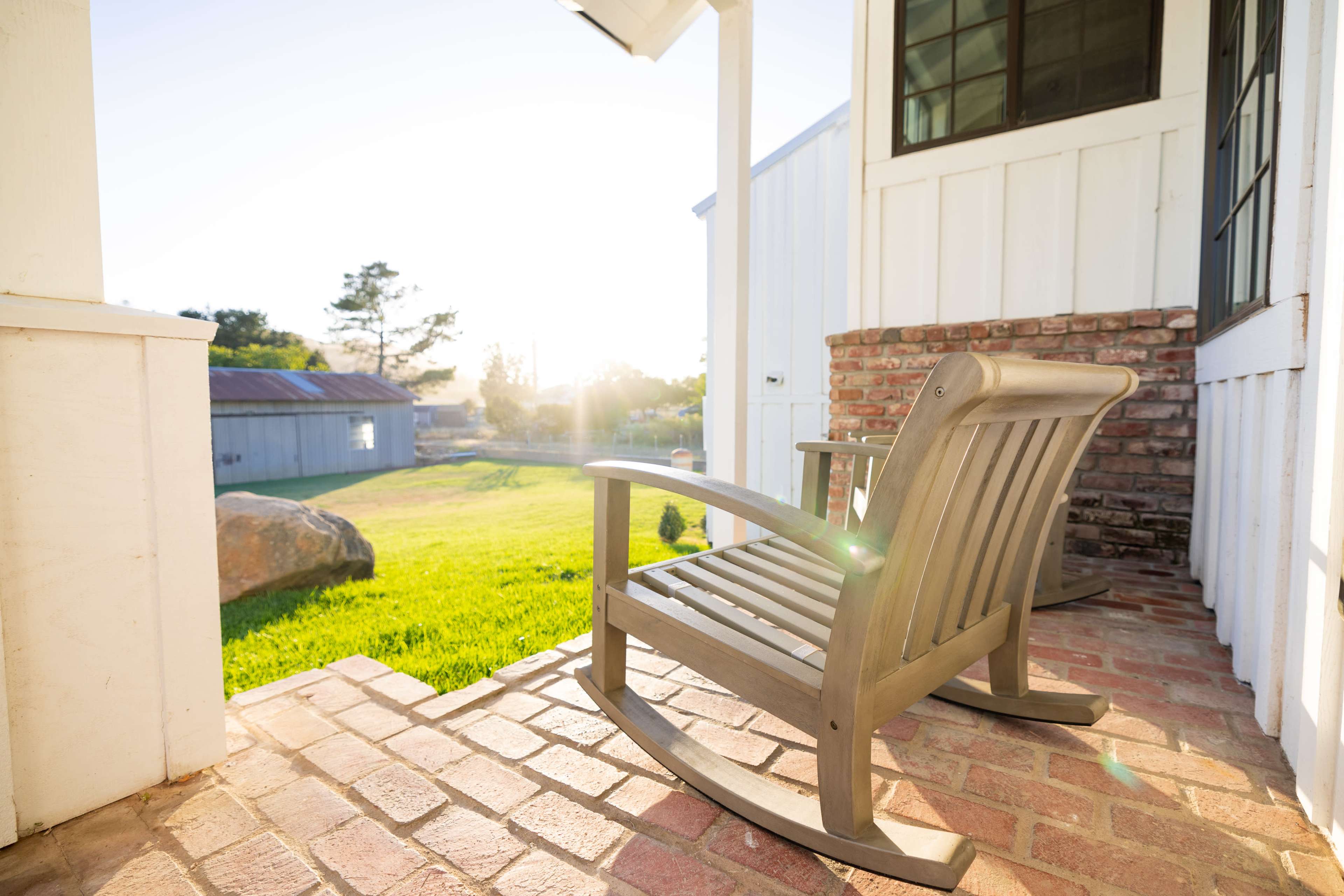 A wooden rocking chair sits on a brick porch overlooking a grassy area and a shed in the background.
