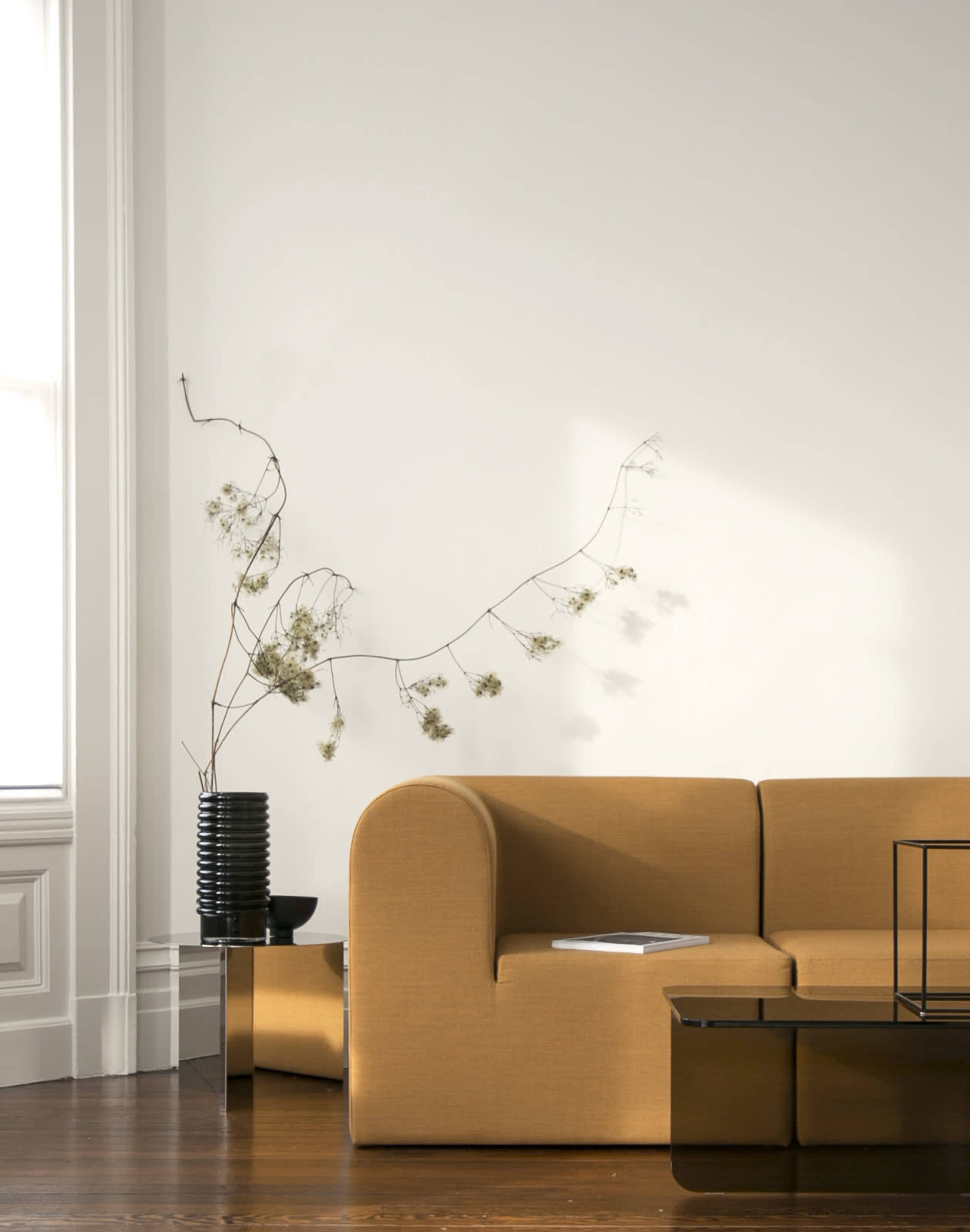 A curved yellow sofa sits against a white wall, next to a black vase with dried flowers and a coffee table.