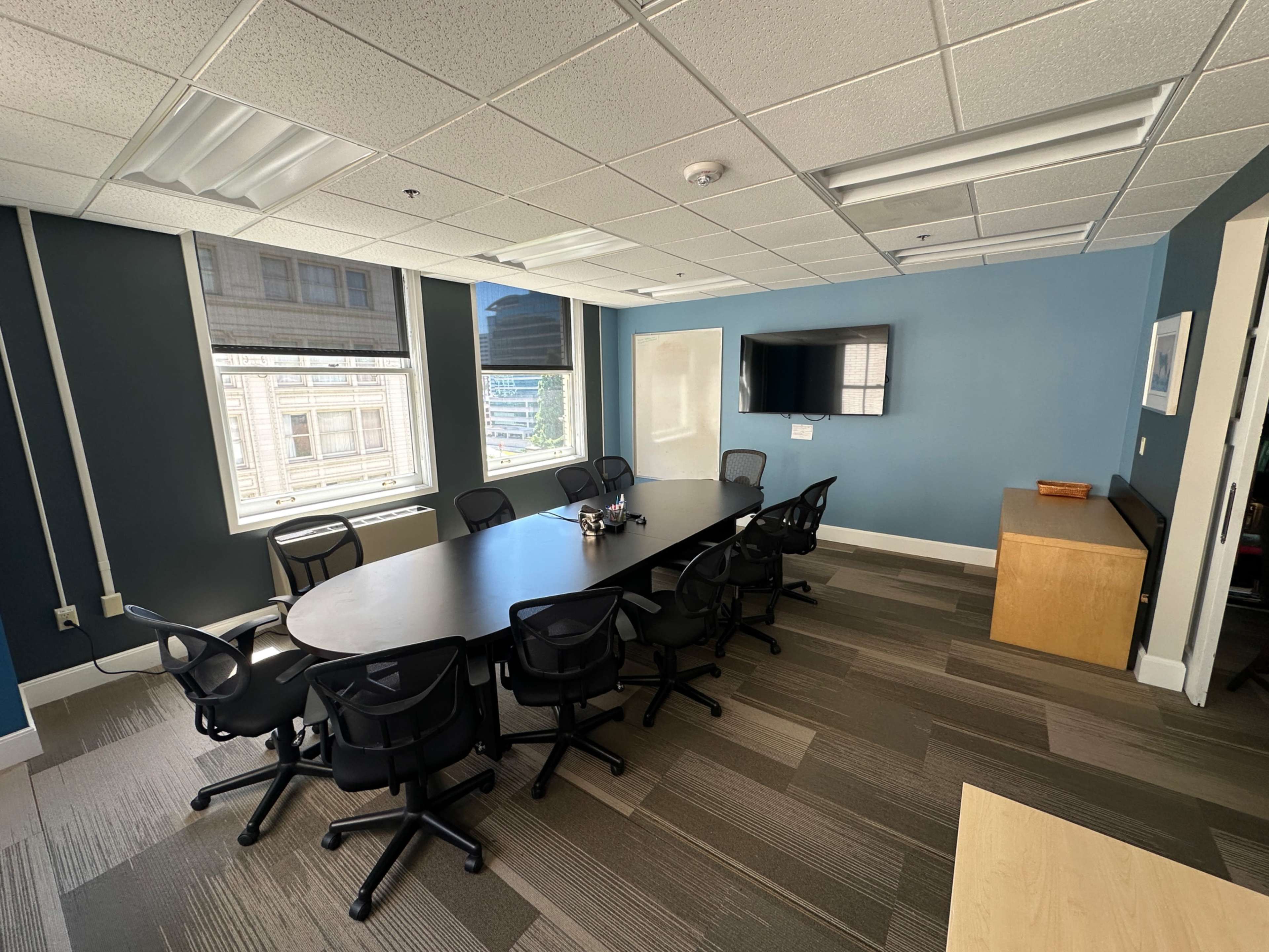 The image shows a modern conference room with a large black table surrounded by swivel chairs, large windows, and a wall-mounted television.