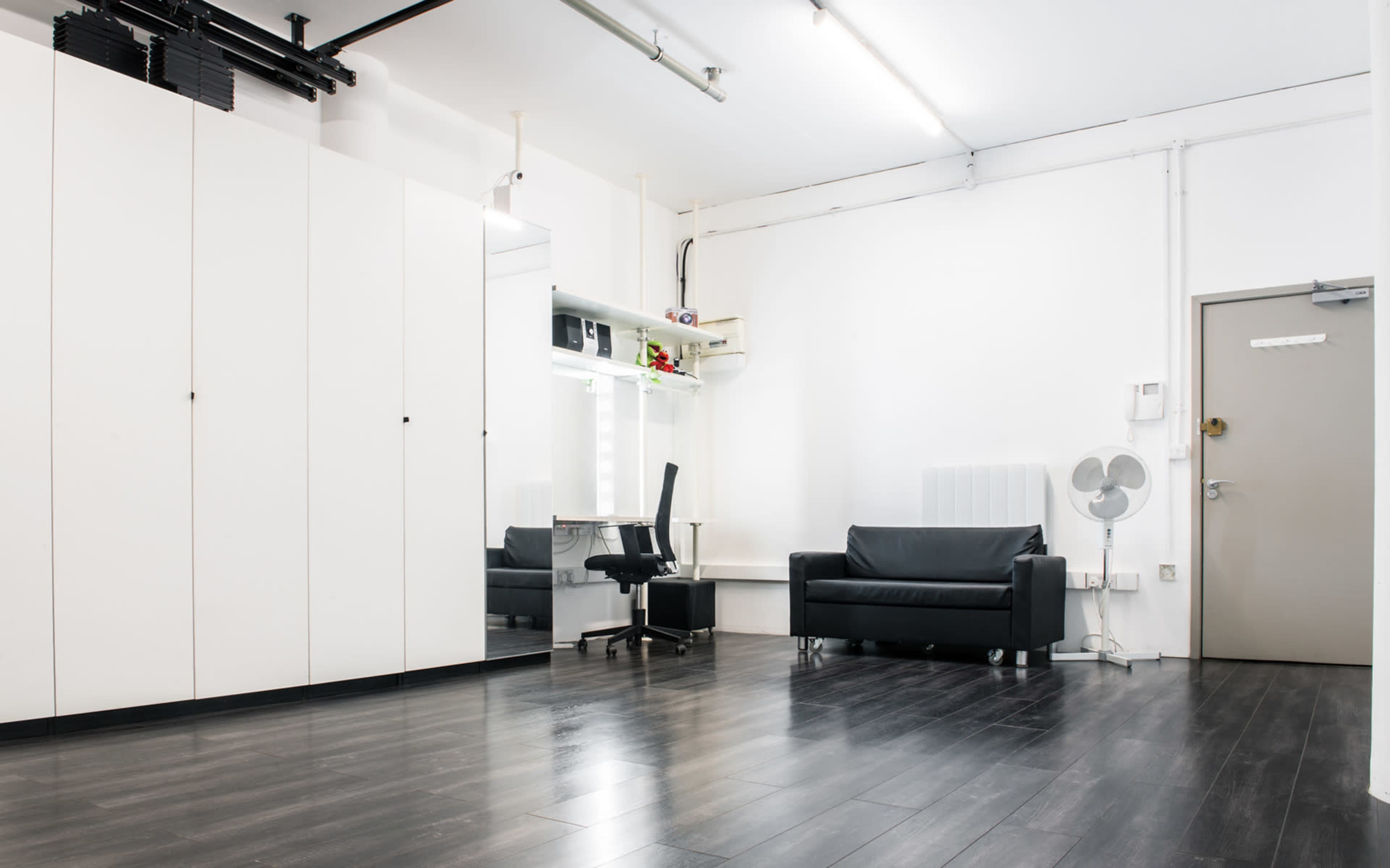 The image shows a minimalist interior space with a black sofa, a desk chair, and built-in white cabinetry.