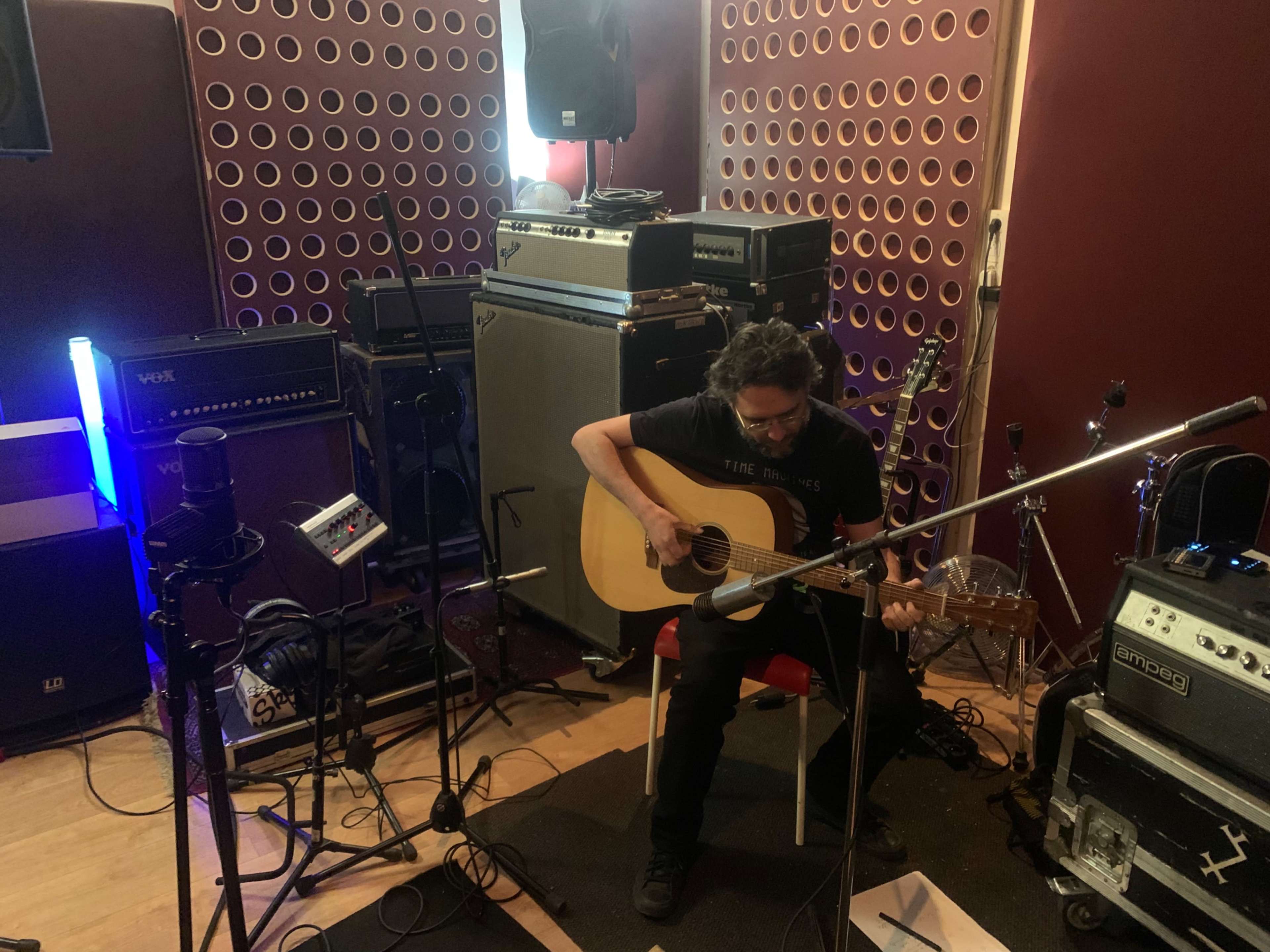 A musician sits on a chair playing an acoustic guitar in a recording studio surrounded by amplifiers and audio equipment.
