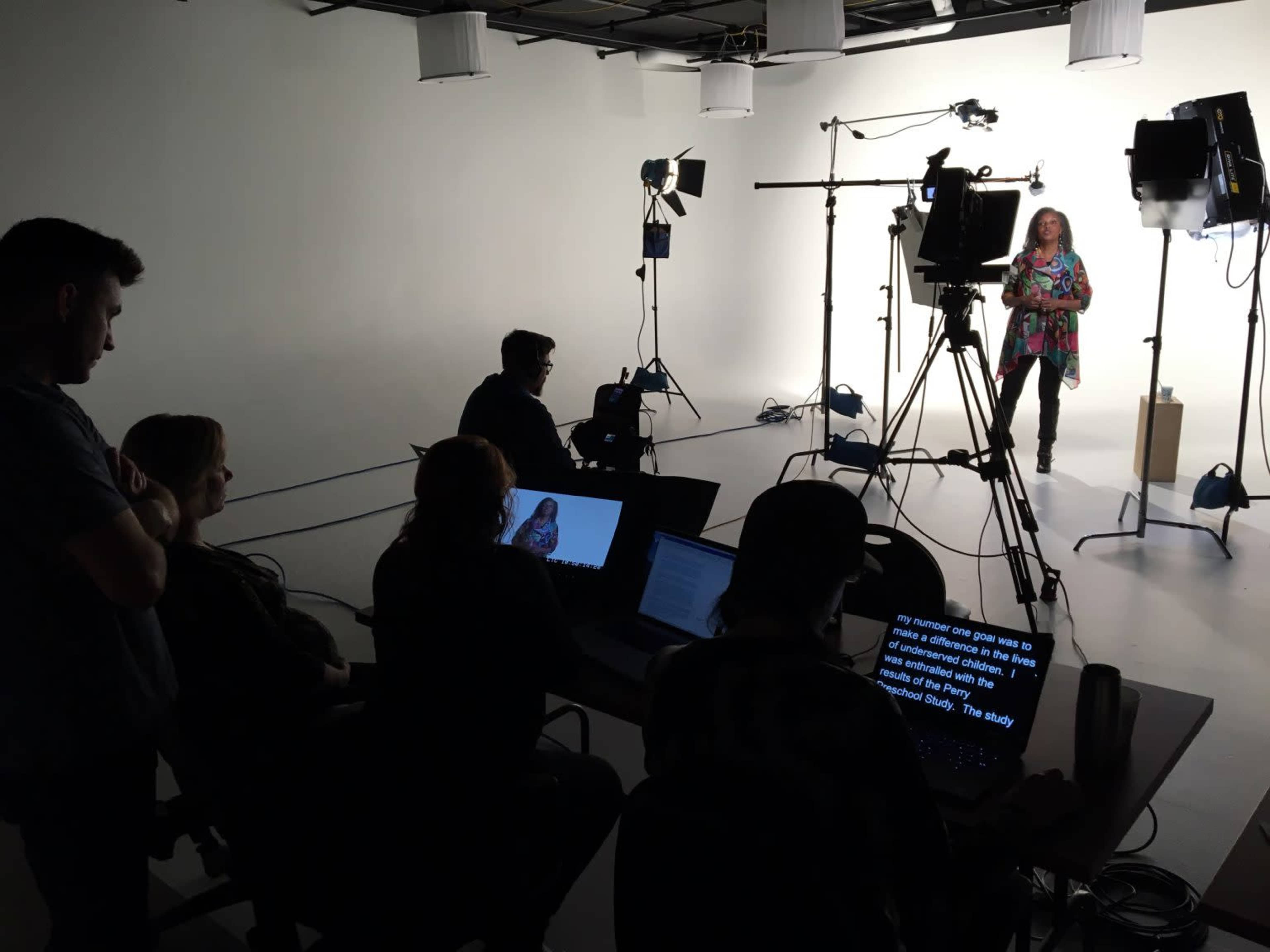 A group of people sits at laptops in a studio while a woman stands in front of a camera under bright lights.