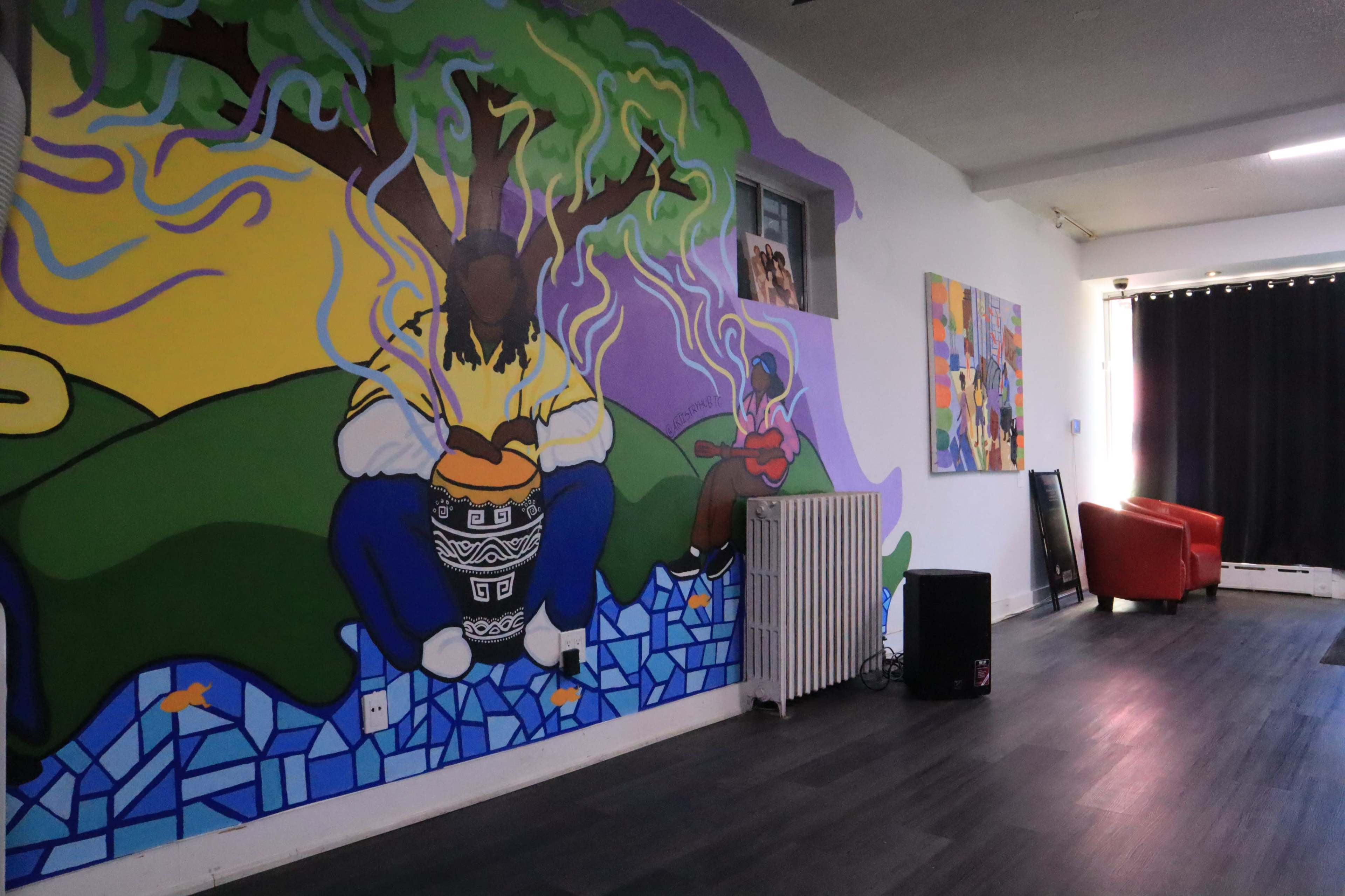 The image shows an interior space featuring vibrant murals on the walls, including a colorful tree and figures, with a radiator and red furniture in the background.