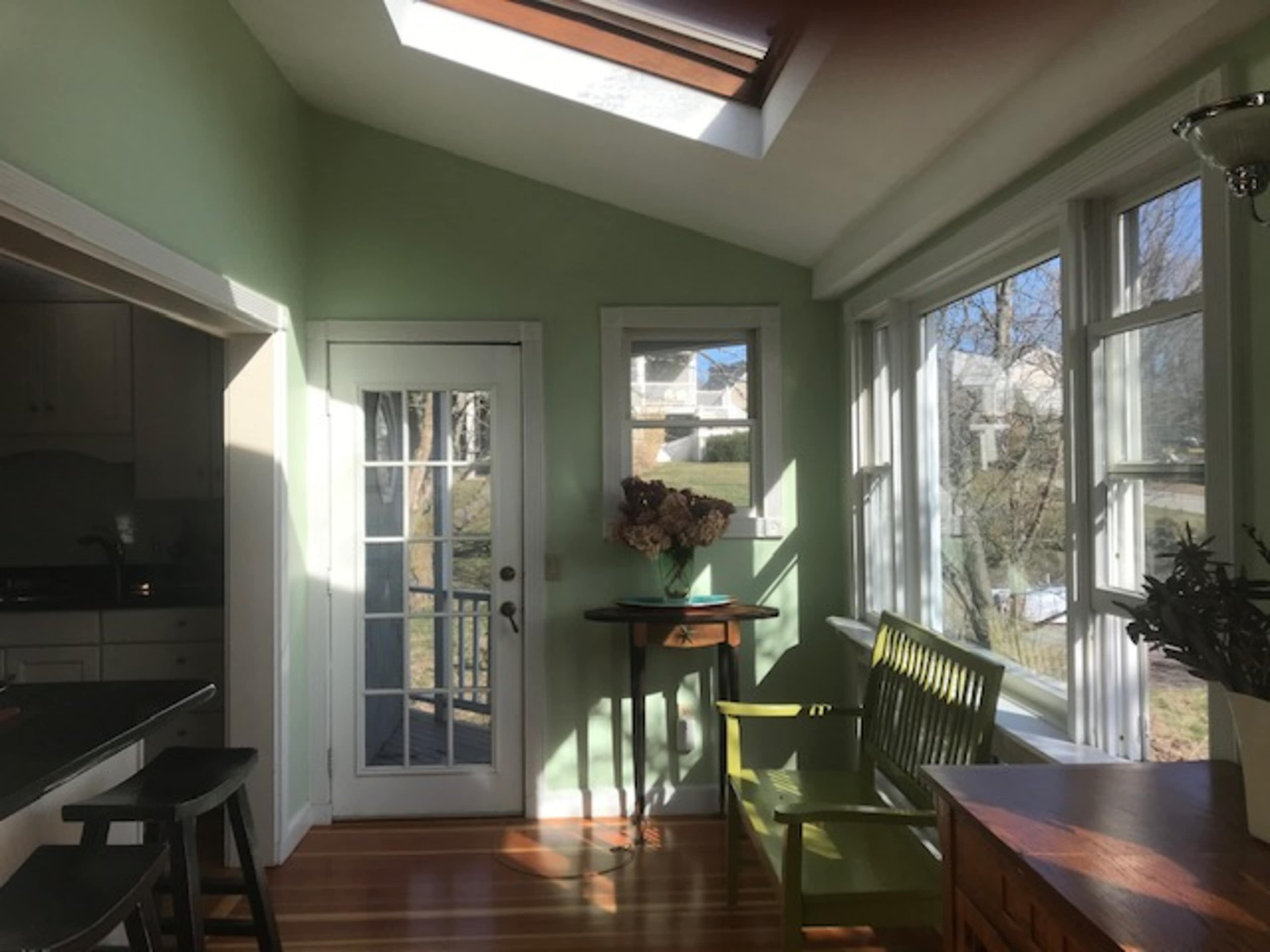 A sunlit room with green walls features a table, a chair, and glass doors leading to a deck.