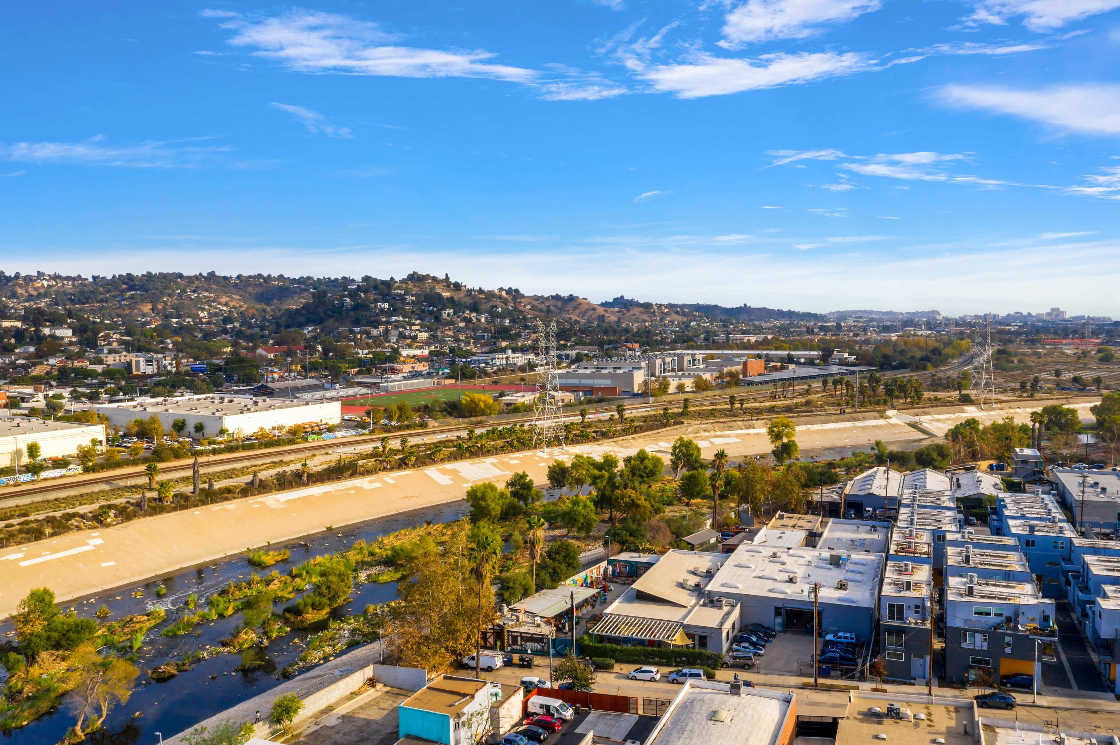 The image shows a landscape with a winding waterway bordered by a road and low vegetation, alongside an industrial area and hills in the background.