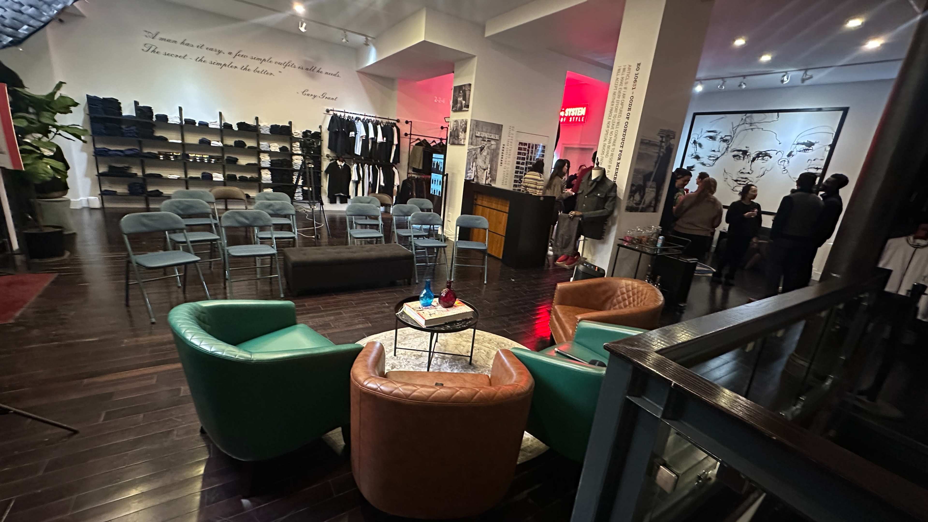 The image shows a modern retail space featuring seating arrangements with a coffee table, racks of clothing, and a gathering of people in the background.