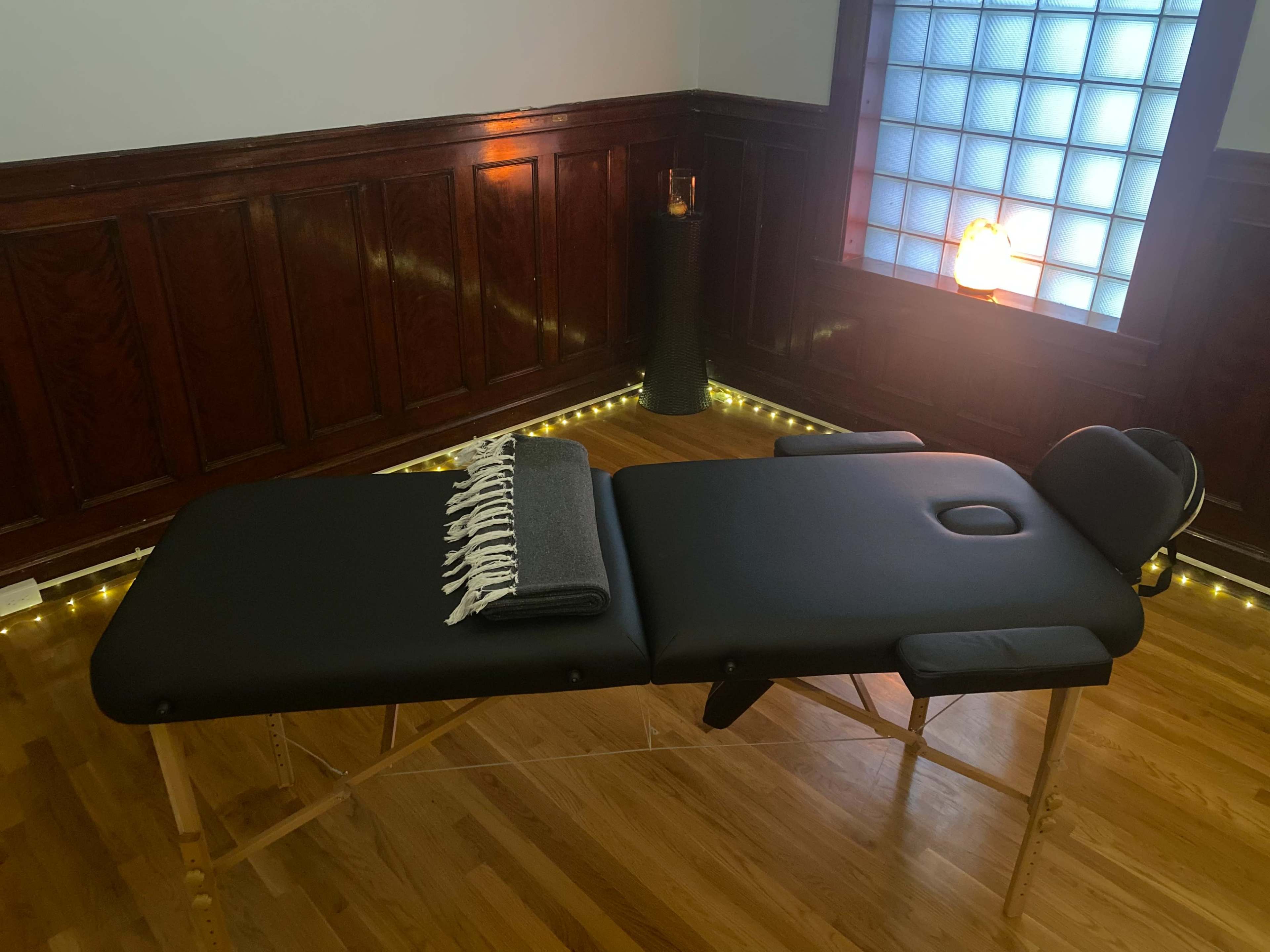 A massage table with a folded blanket is positioned in a softly lit room featuring wooden walls and a frosted glass window.