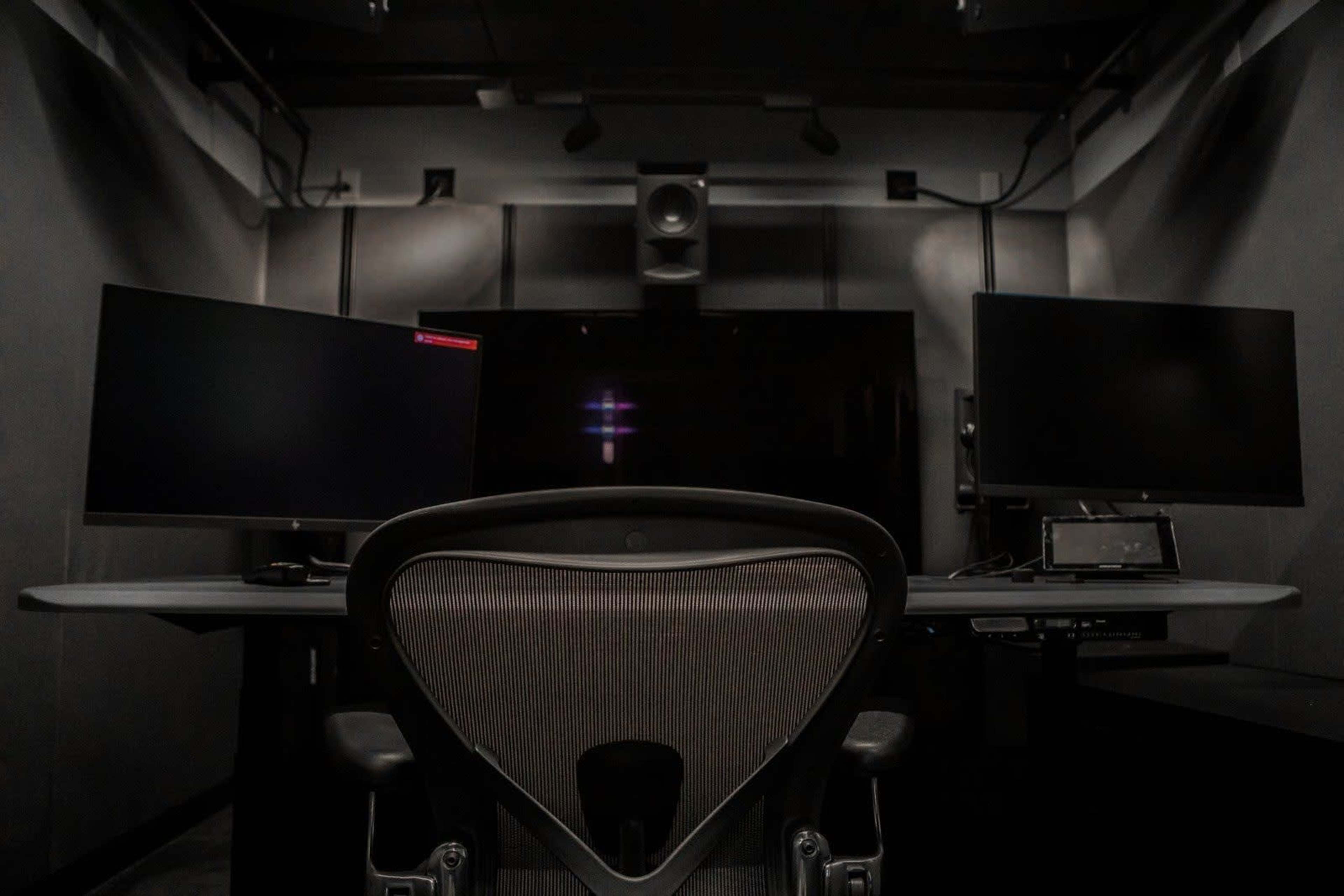 A modern workspace features two computer monitors and a chair facing a large screen in a dimly lit room.