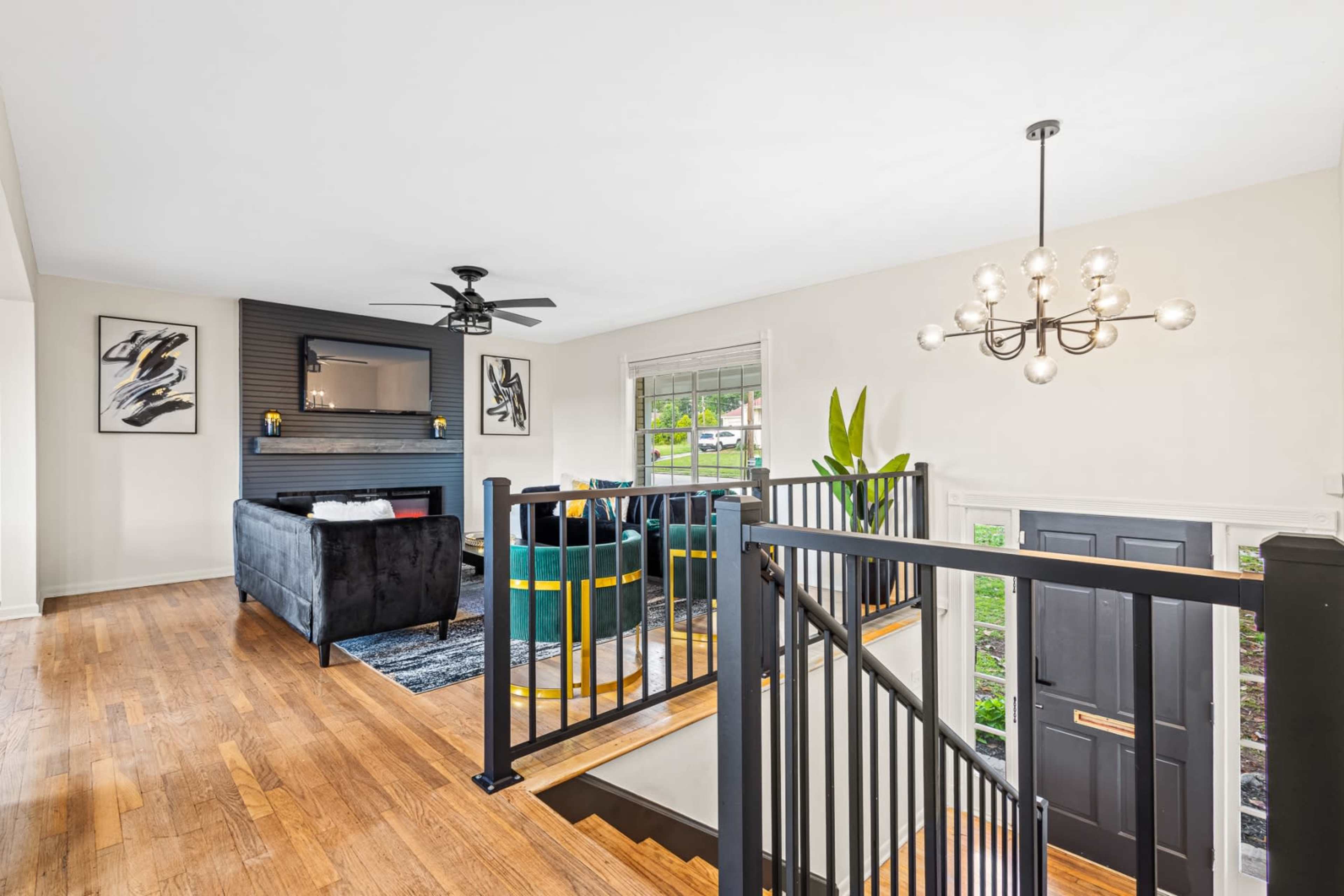 The image displays a modern living room with a black couch, a ceiling fan, and a staircase leading to an entrance area.