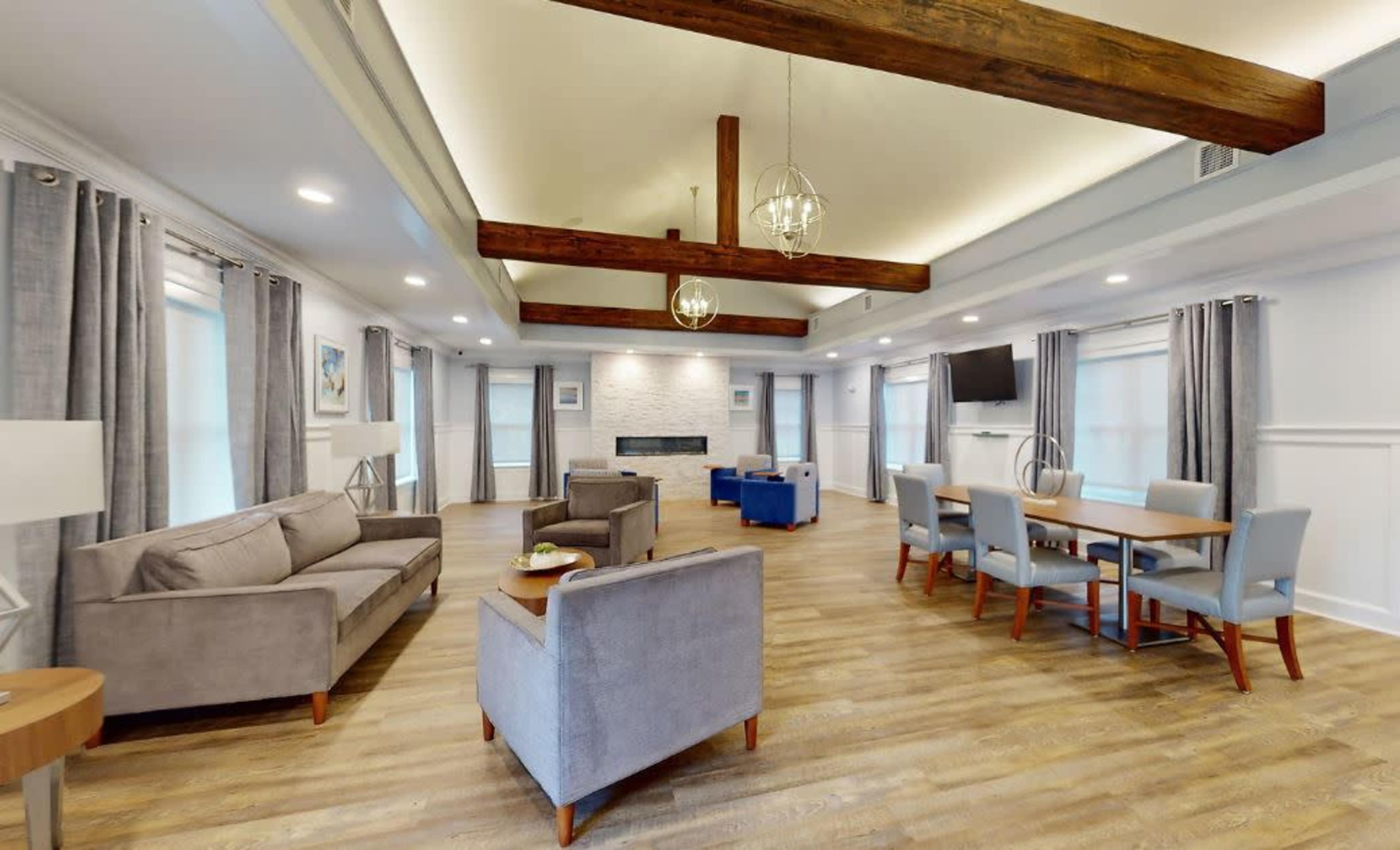 The image shows a spacious living area with a seating arrangement, a dining table, a modern fireplace, and large windows, all featuring wooden beams across the ceiling.