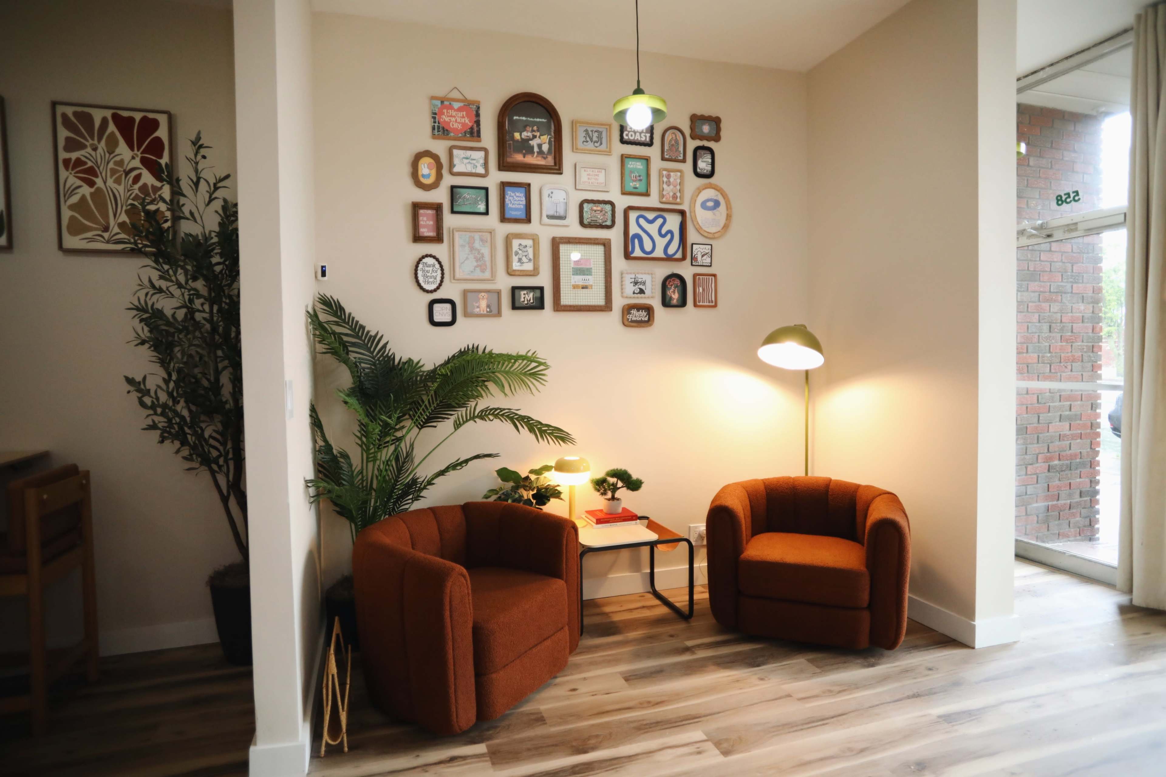 The image depicts a cozy corner with two orange armchairs, a small table, and a gallery wall adorned with various framed artworks, next to a potted plant and a standing lamp.