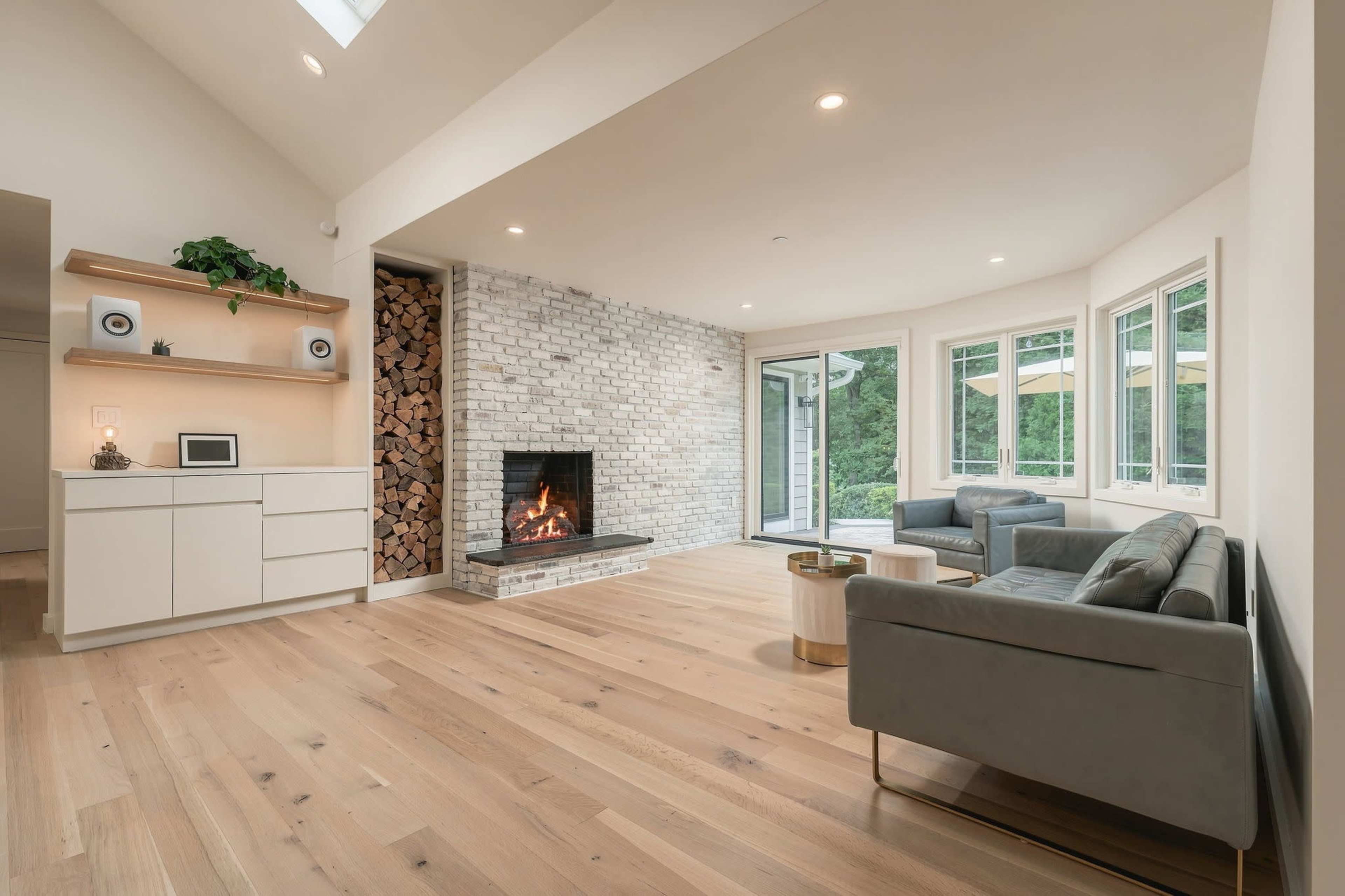 A modern living room features a brick fireplace, large windows, and light wood flooring.