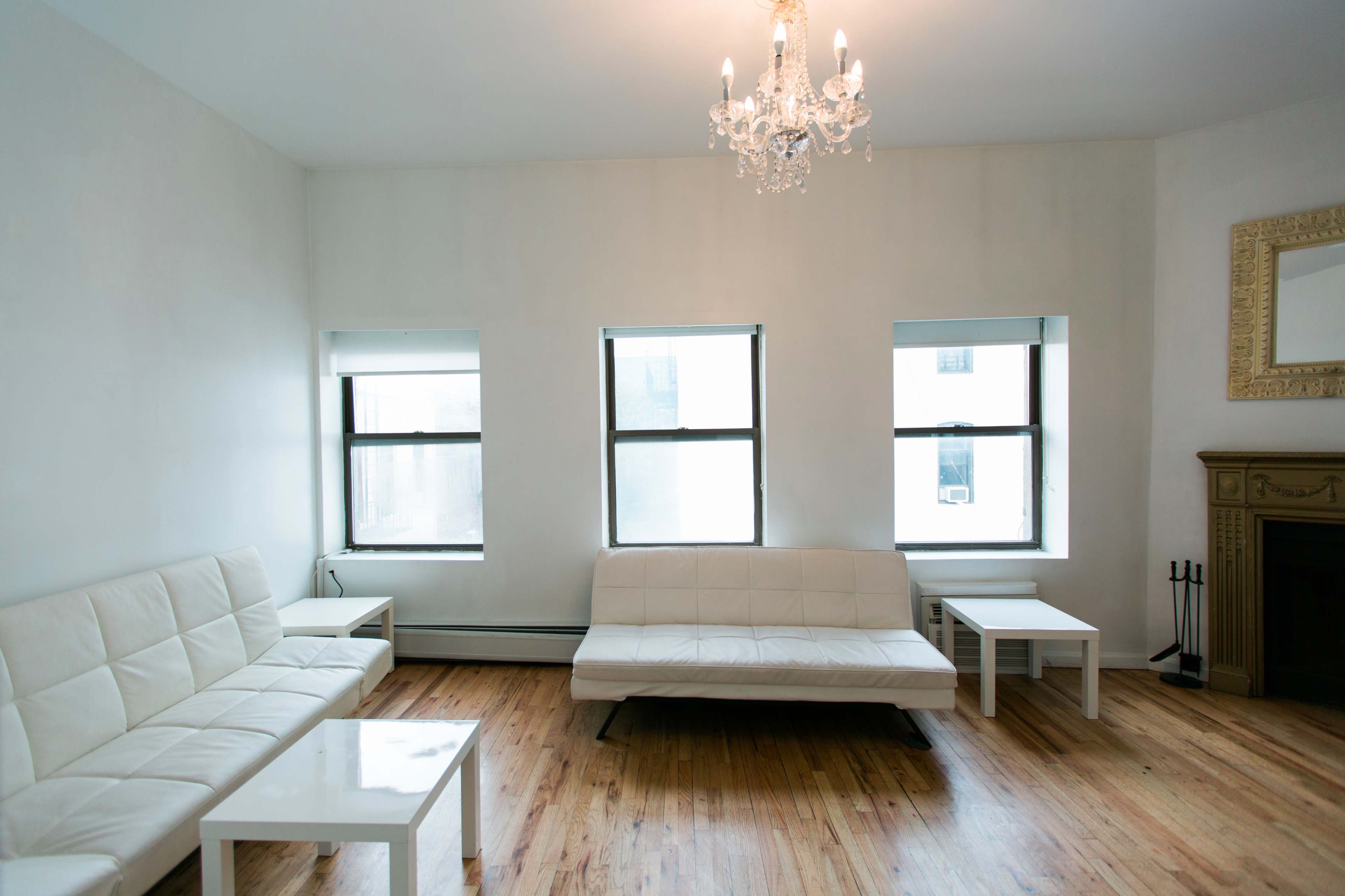 A modern living room features a white leather sofa and coffee table, three windows, and a chandelier hanging from the ceiling.