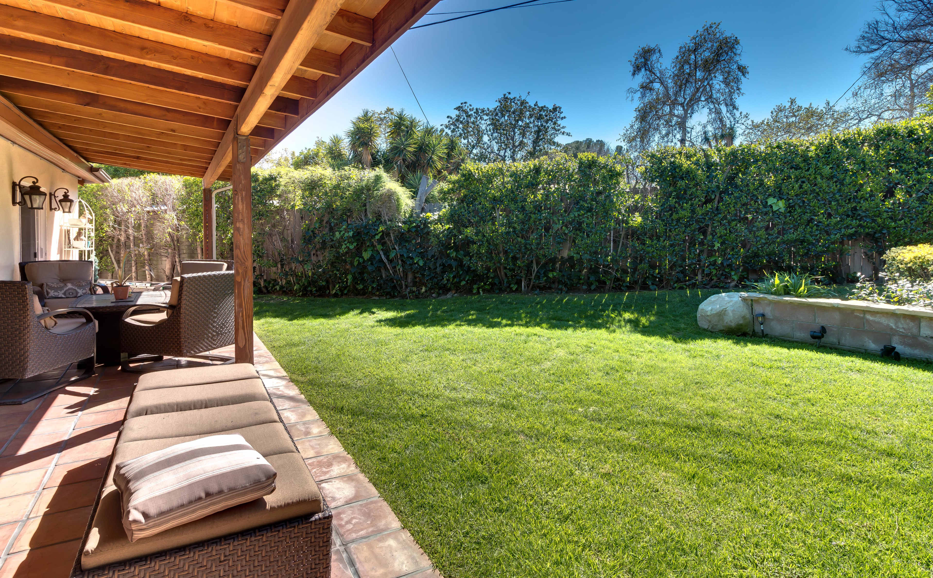 A well-maintained backyard features a grassy lawn, patio seating under a wooden overhang, and dense greenery in the background.