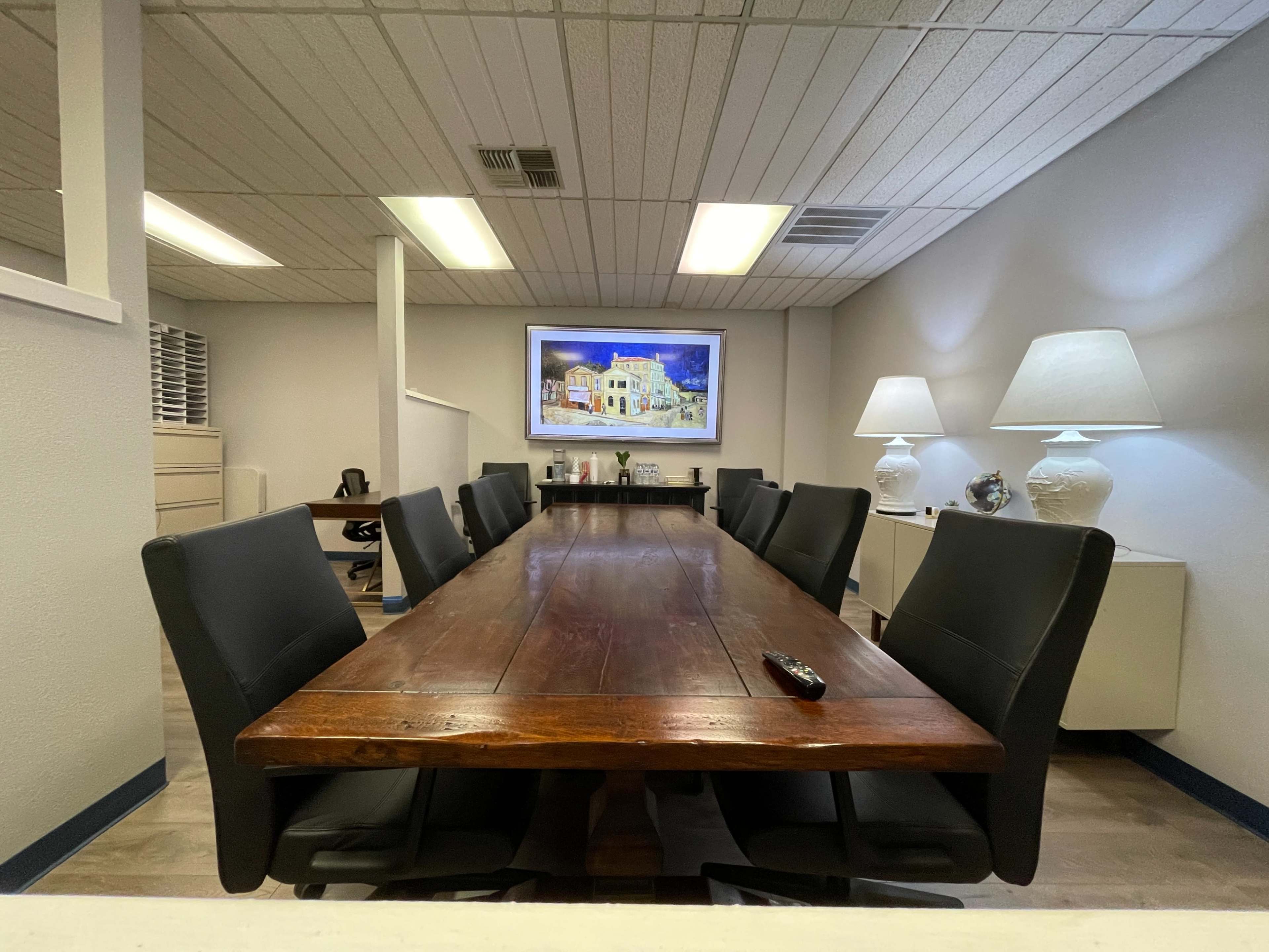 A long wooden conference table is surrounded by black chairs in a well-lit office meeting room featuring a large artwork on the wall.