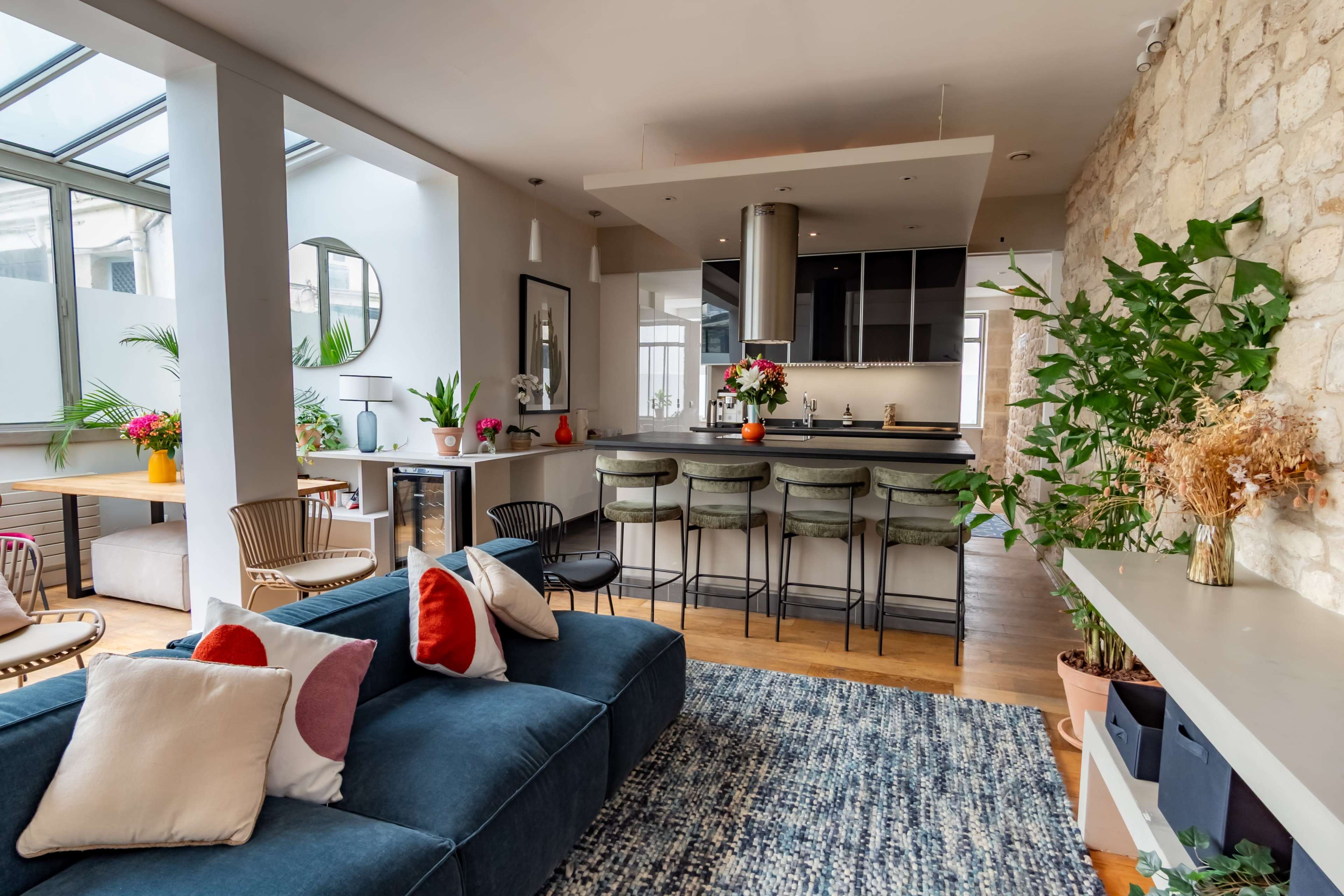 The image shows a modern apartment interior featuring a living area with a blue sofa, a kitchen with bar seating, and a mix of plants and decorative items.