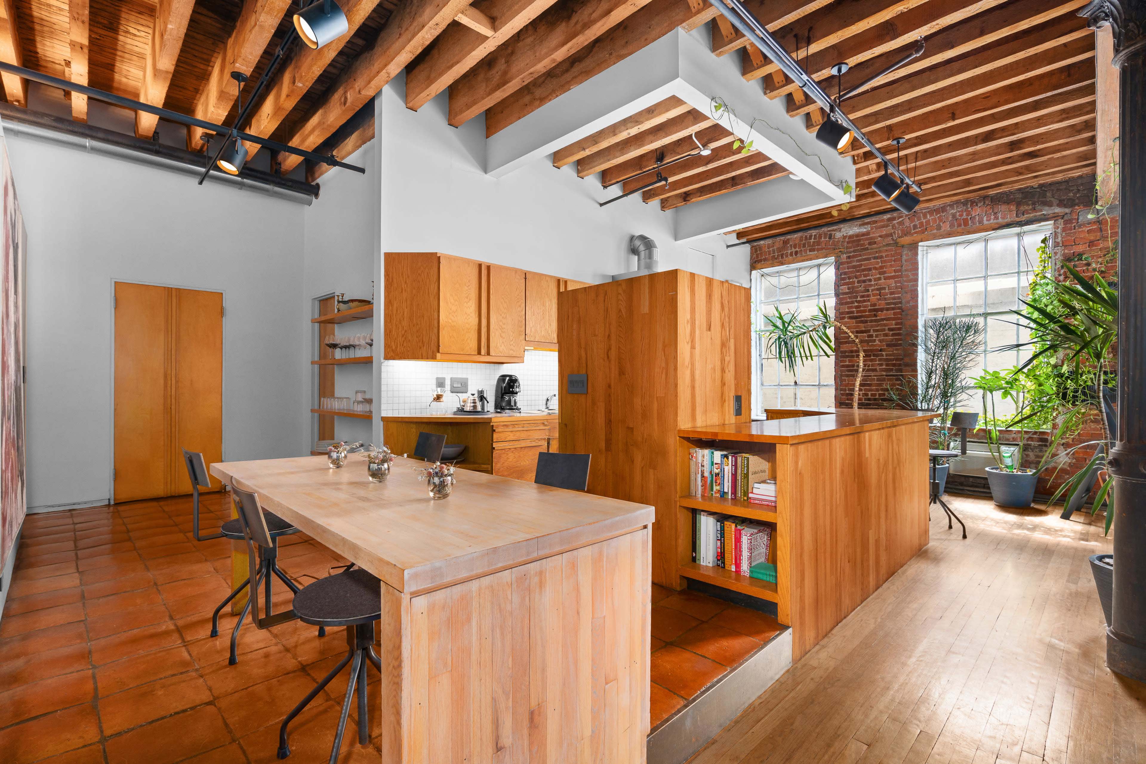 The image shows a modern kitchen and dining area with wooden beams, a long wooden table, and built-in shelving against a backdrop of large windows and potted plants.