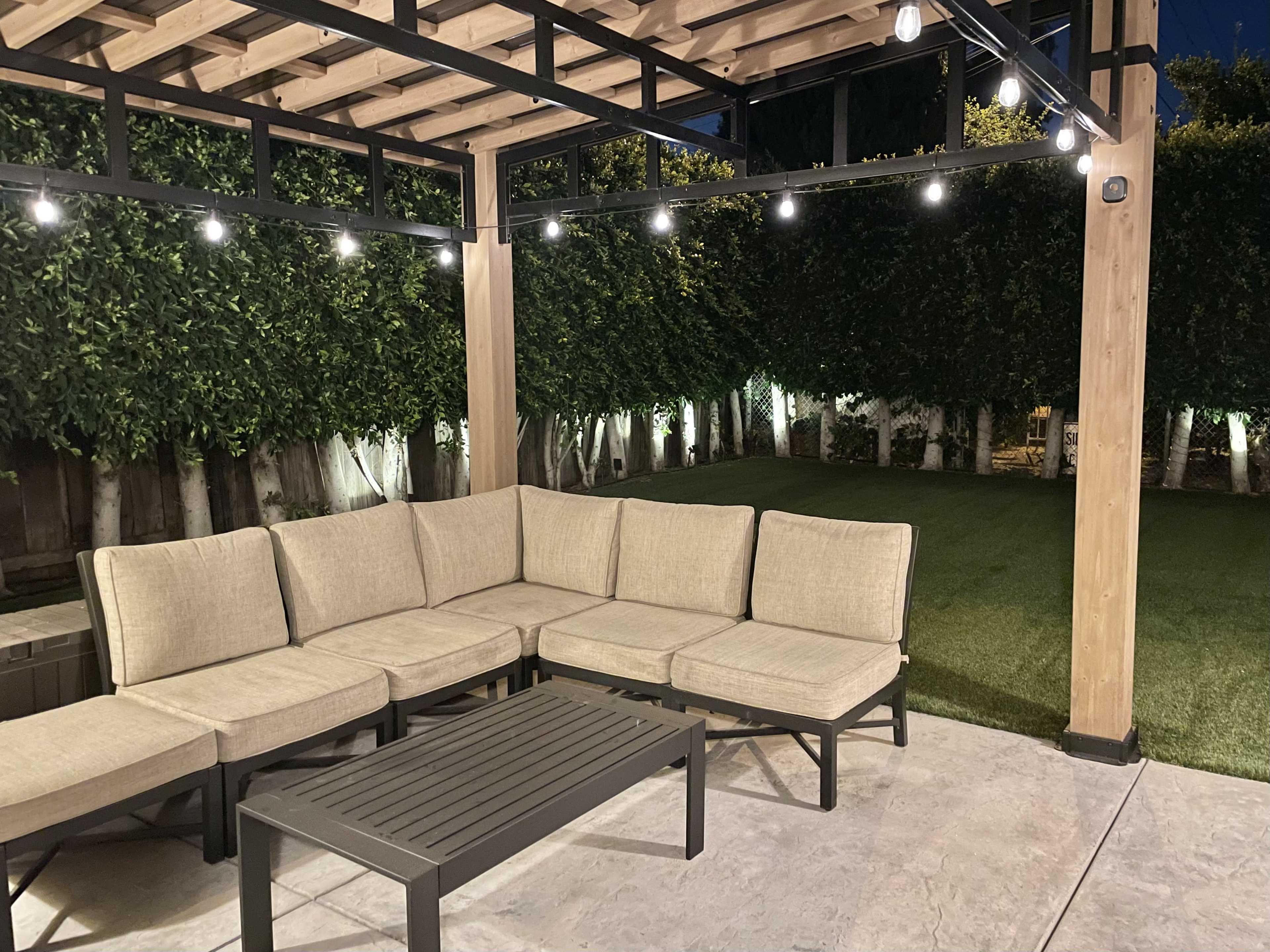 A modern outdoor seating area features a sectional sofa and a coffee table under a pergola with string lights, surrounded by manicured greenery.