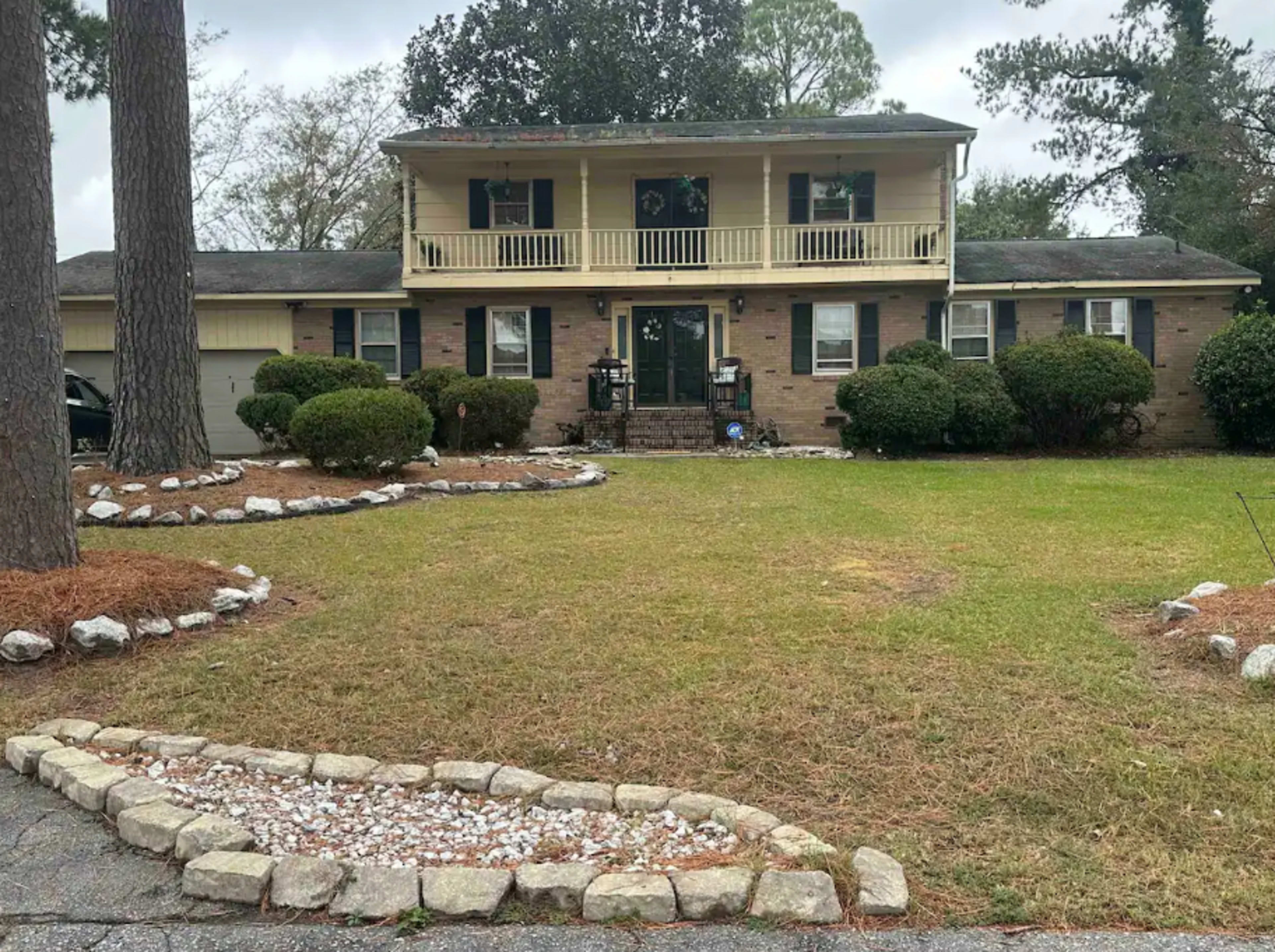 A two-story house with a front porch, flanked by trees and a landscaped yard featuring a rock-bordered flower bed.