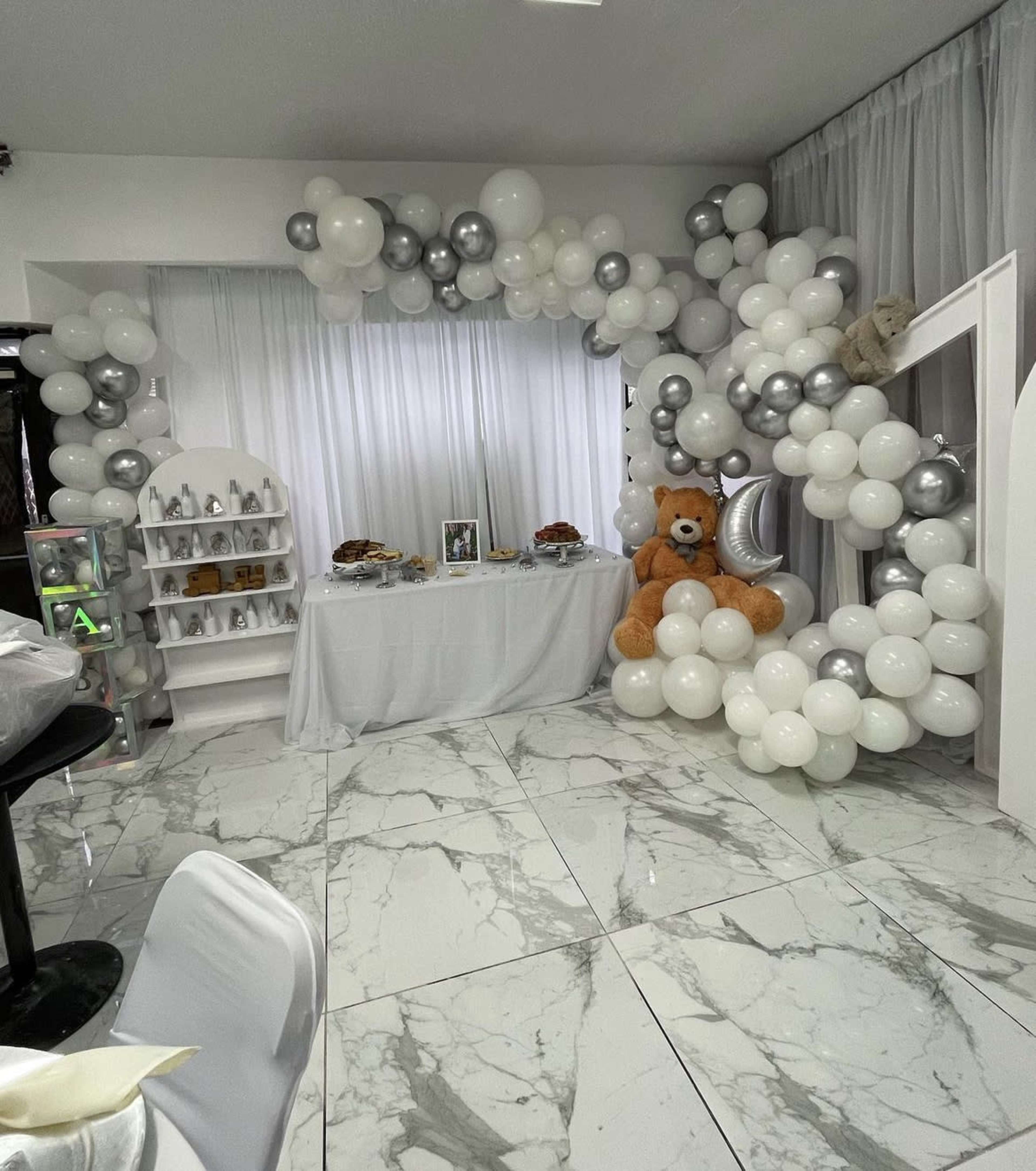 The image shows a decorated event space featuring a backdrop of white and silver balloons alongside a table with snacks and a large teddy bear.