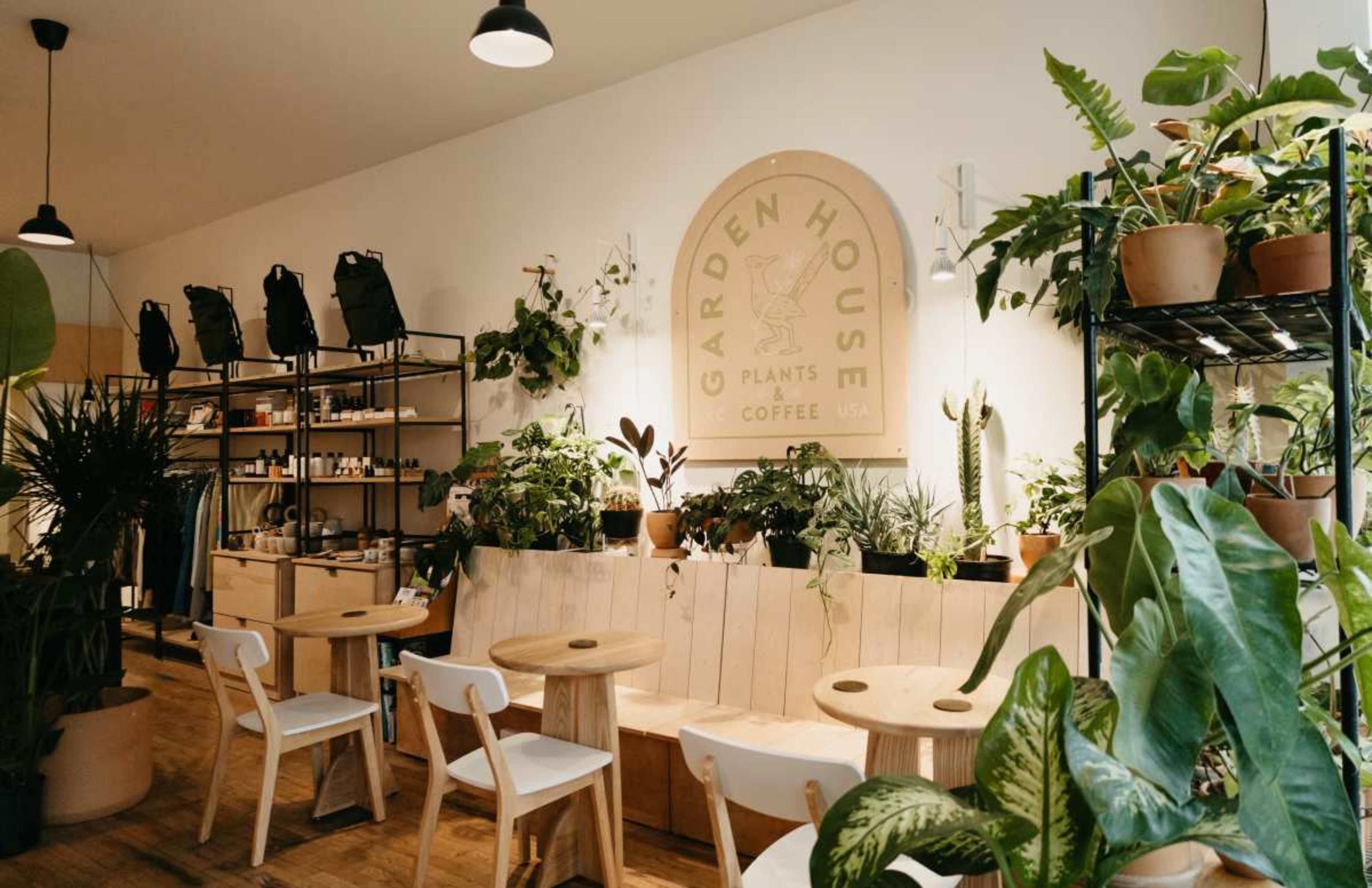 The image shows a cozy indoor space filled with various potted plants, wooden seating, and a sign that reads "GARDEN HOUSE" while displaying coffee and plant-related items for sale.