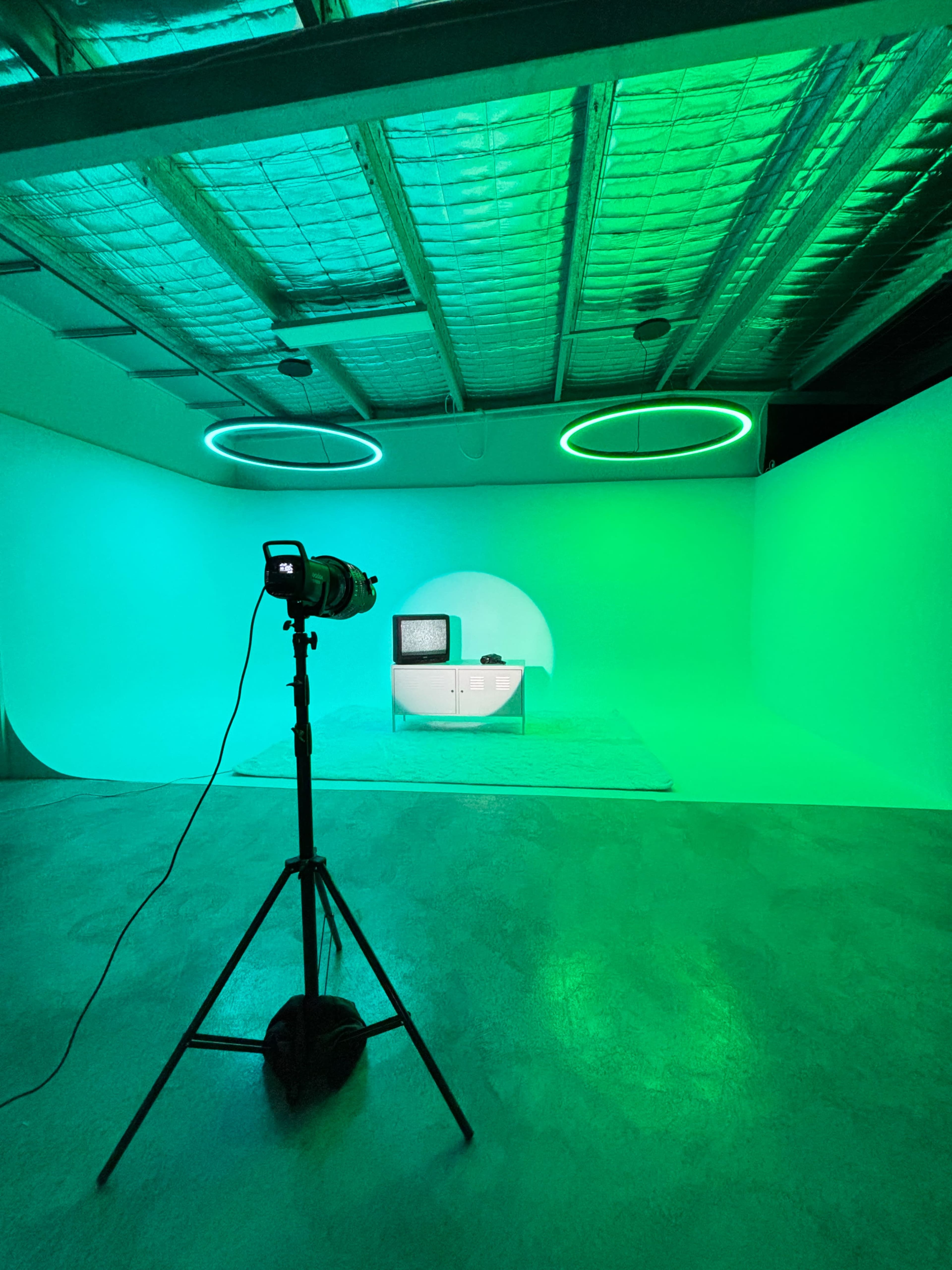 A camera on a tripod is positioned in a brightly lit studio featuring green lighting, with a vintage television set placed on a low table against a blank backdrop.