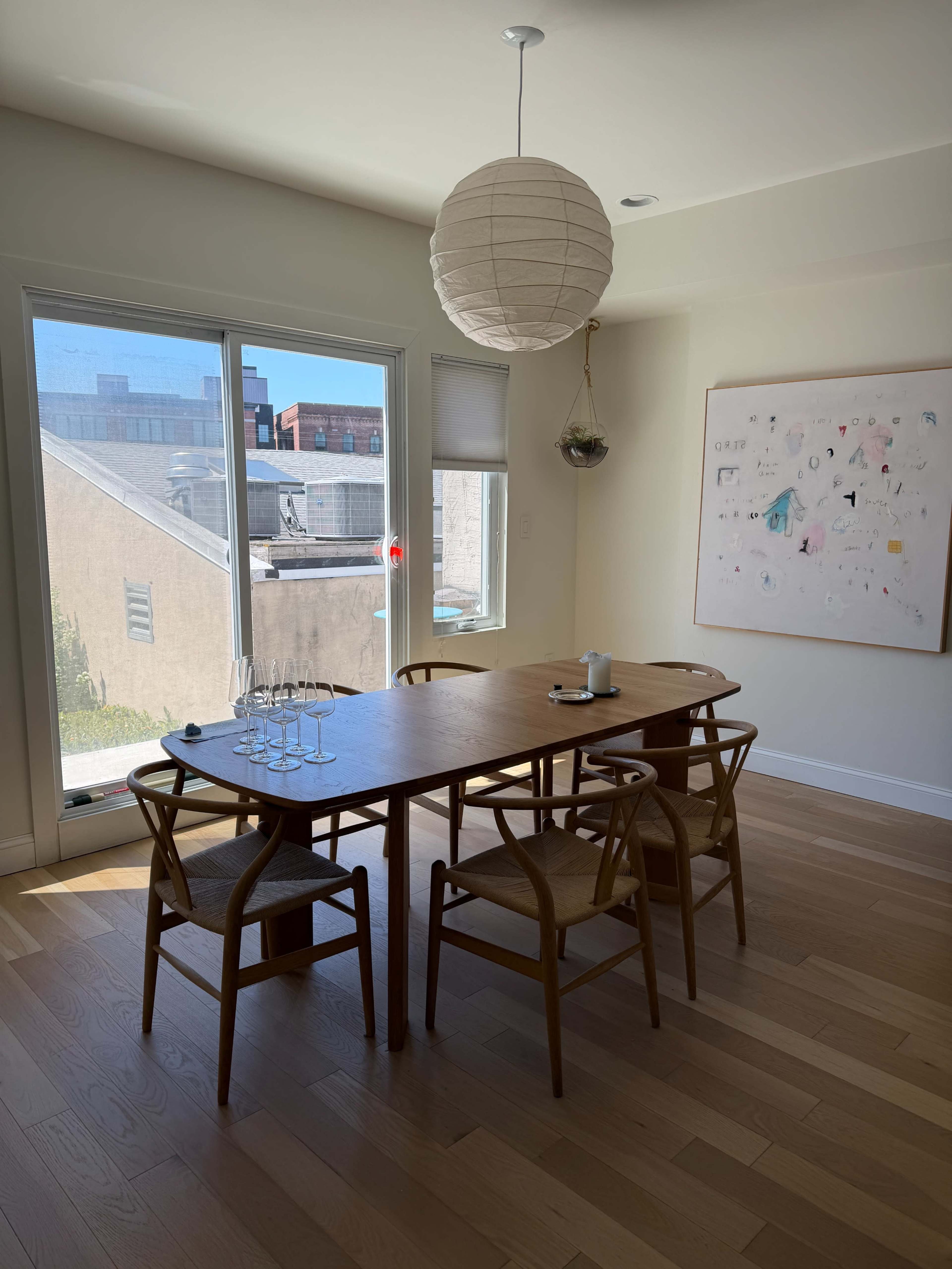 A wooden dining table with four chairs is set near a large window, featuring a pendant light above and a piece of art on the wall.