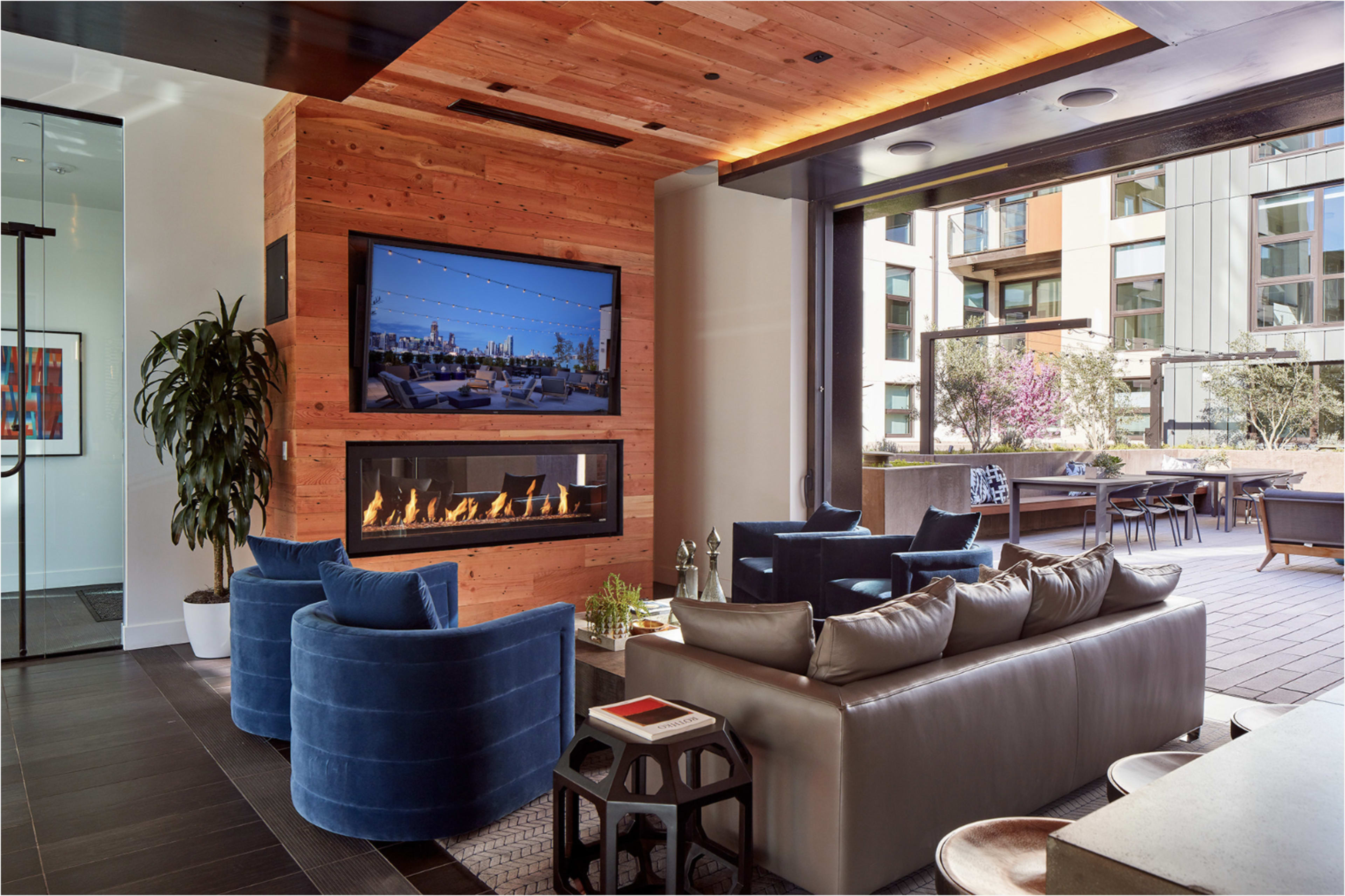 The image shows a modern lounge area featuring a wooden accent wall, a large screen TV, plush seating, and a gas fireplace, all set in an open space with large windows overlooking an outdoor patio.